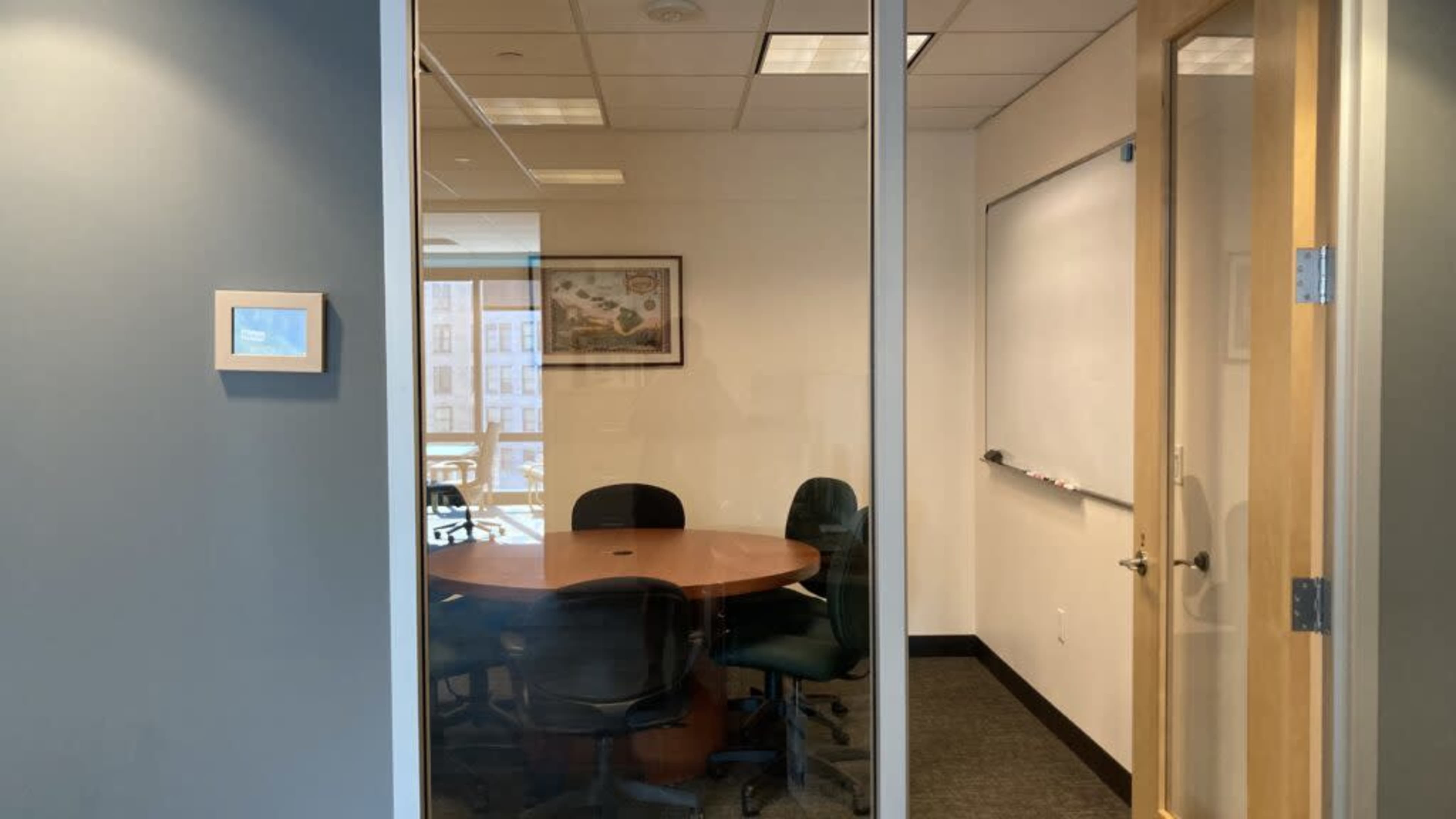 The image shows a conference room with a round wooden table surrounded by chairs, a whiteboard on the wall, and a window allowing light to enter.
