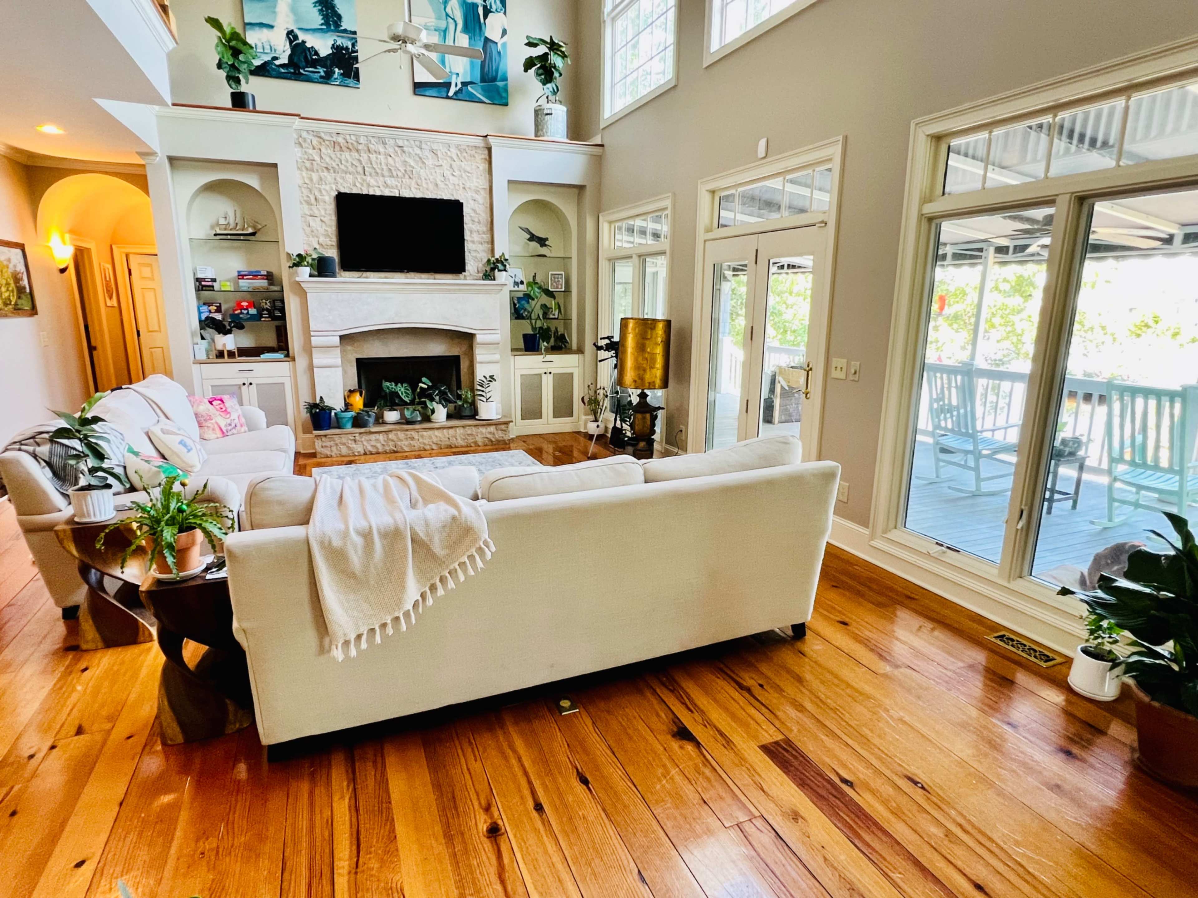A spacious living room features a large white sofa, wooden flooring, and a stone fireplace, with windows that overlook a deck.