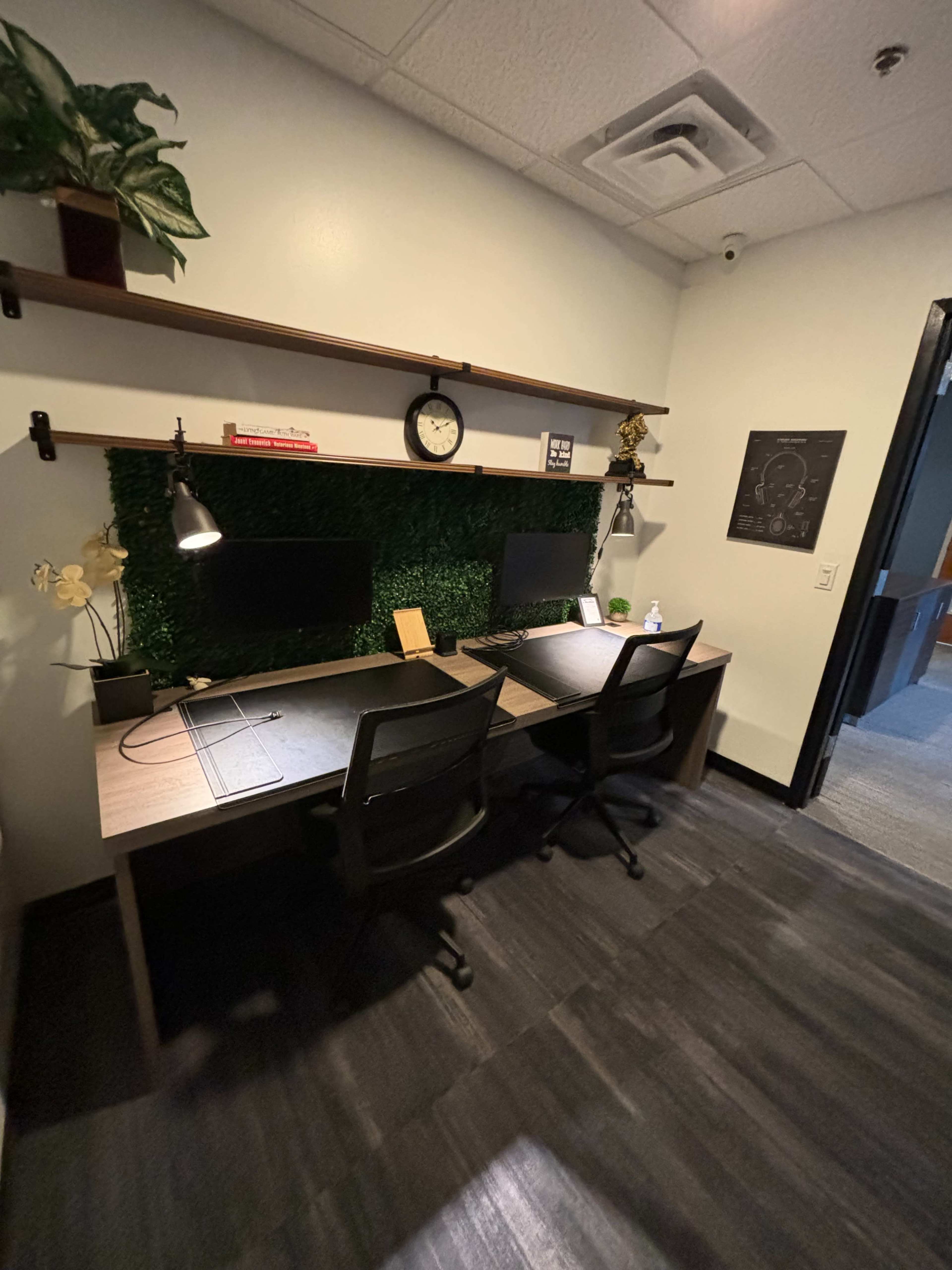 A modern office workspace featuring two desks with monitors, a clock, and a green wall decor made of plants.