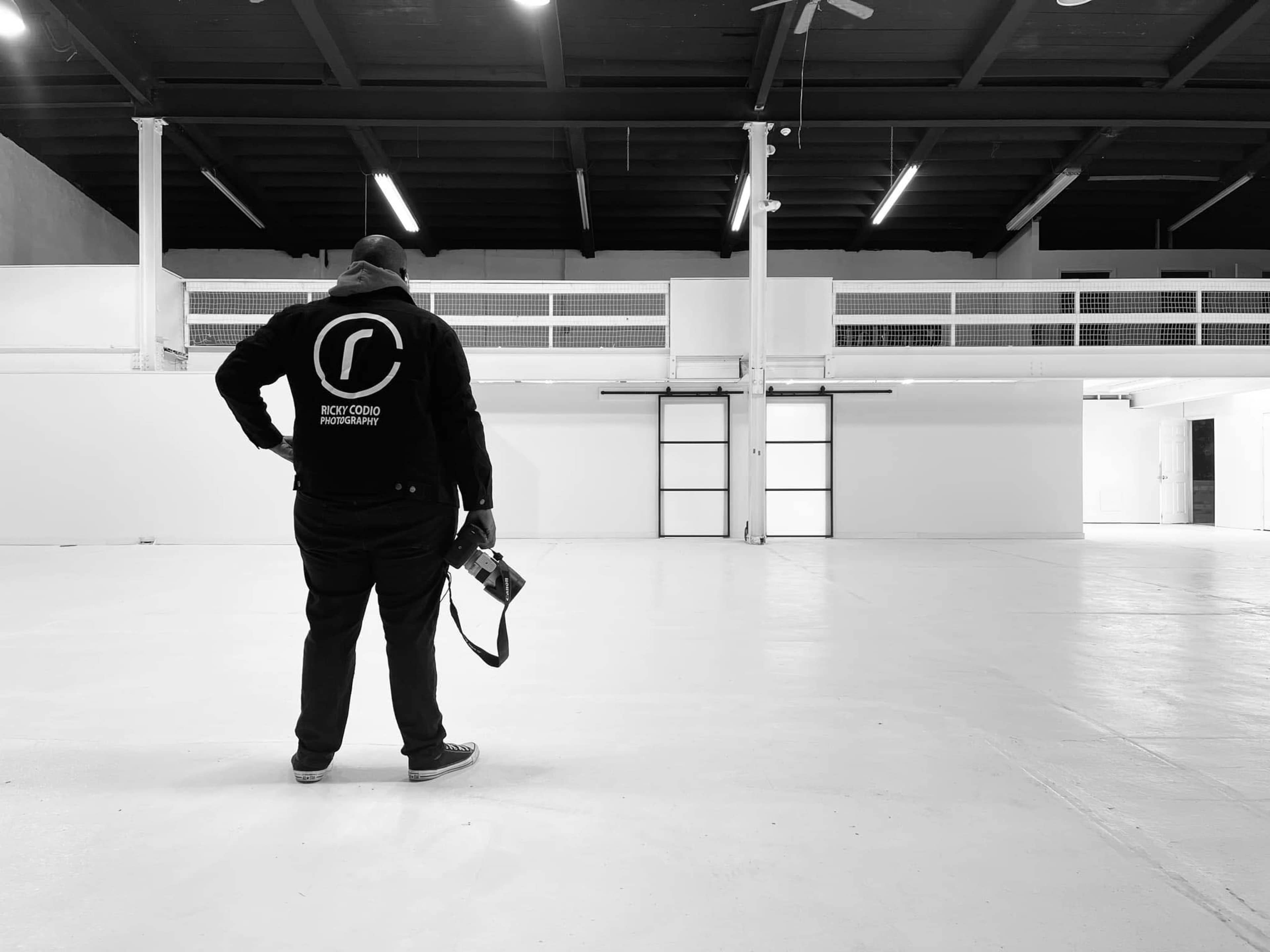 A person stands with a camera in an empty, spacious industrial-style room with white walls and a high ceiling.