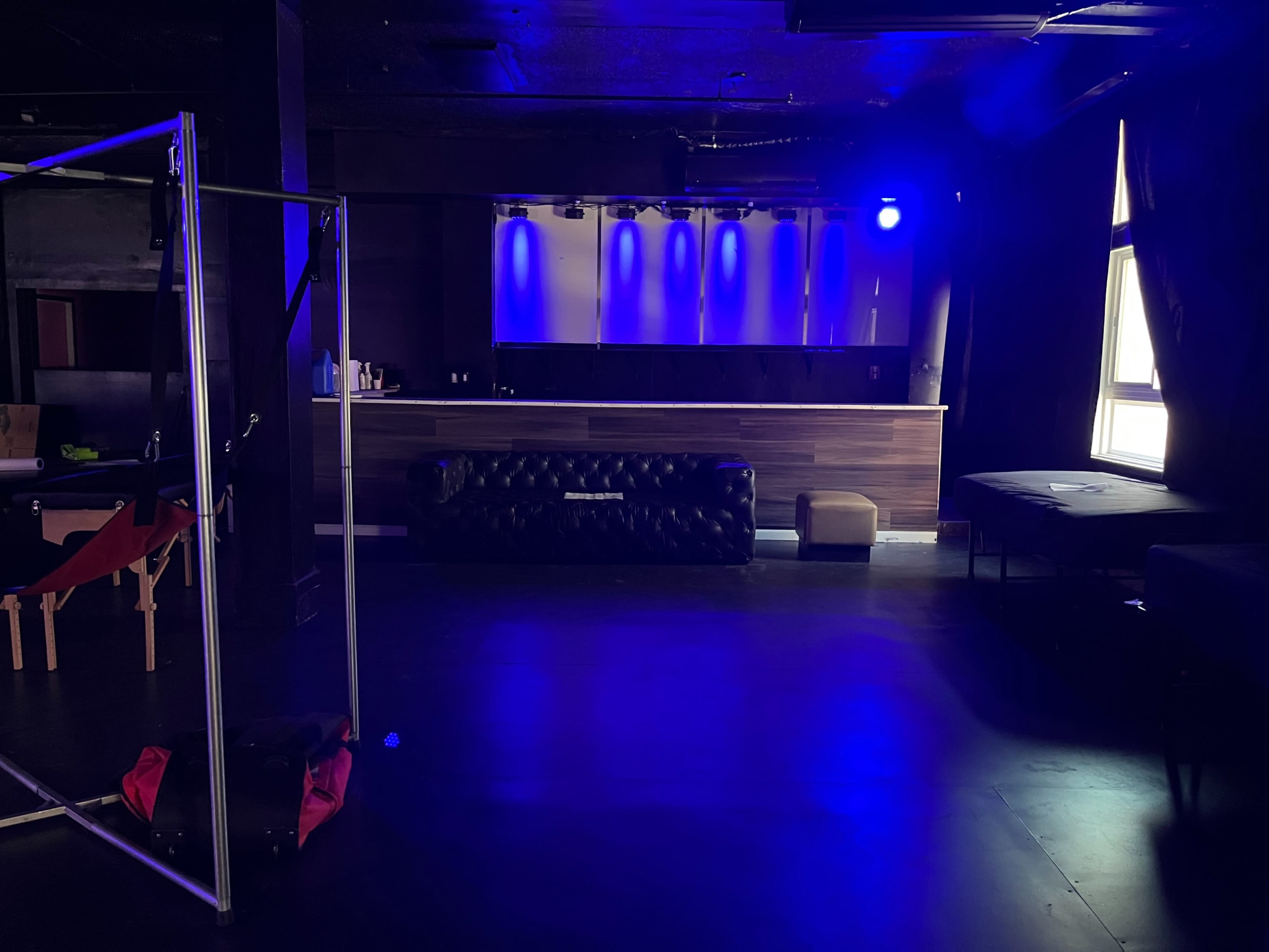 A dimly lit lounge area with a black sofa, a stage with blue lighting, and scattered furniture against a dark backdrop.