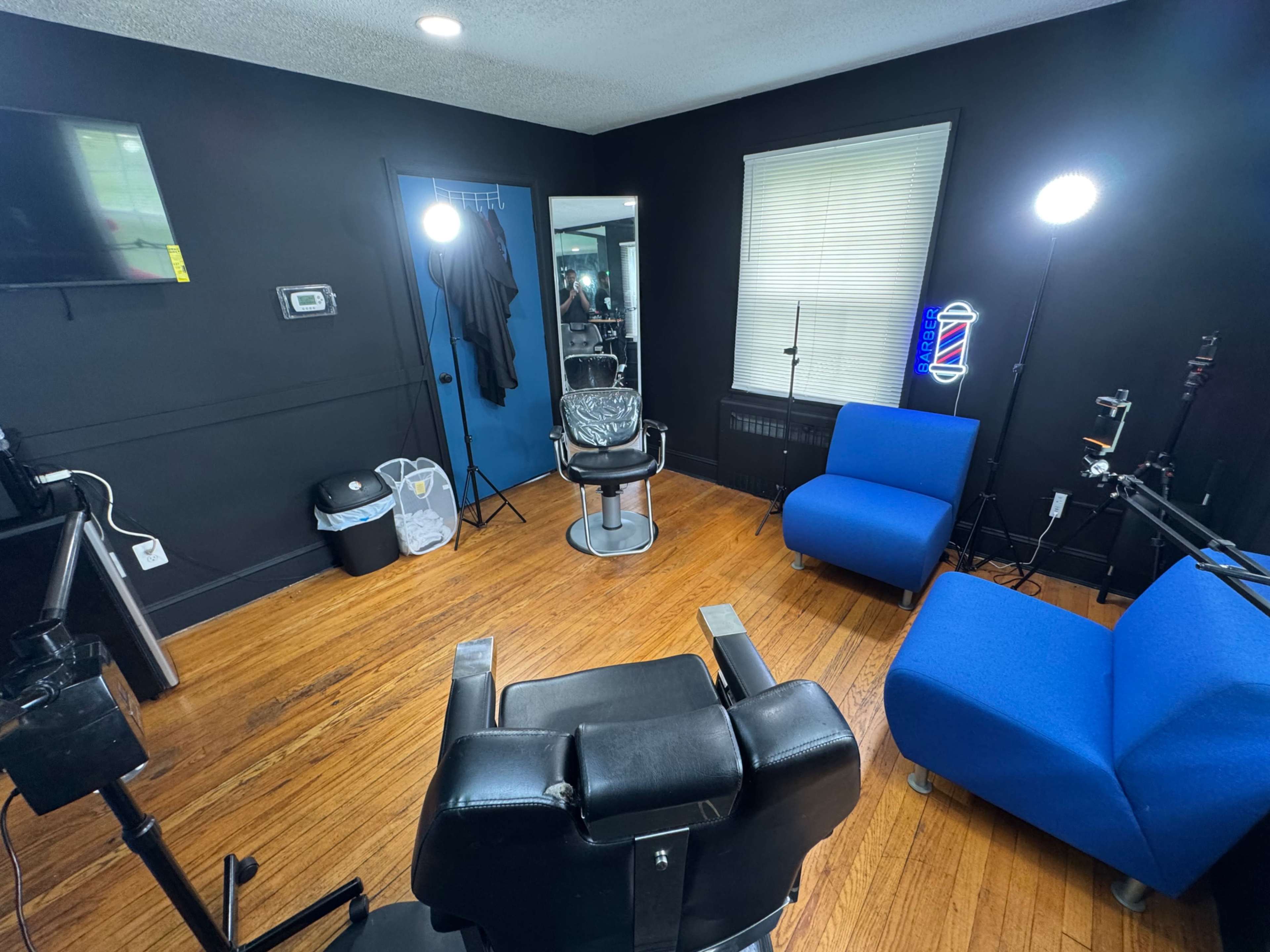 The image shows a barber shop interior with a black wall, a barber chair in the center, two blue chairs, a mirror, and studio lighting.