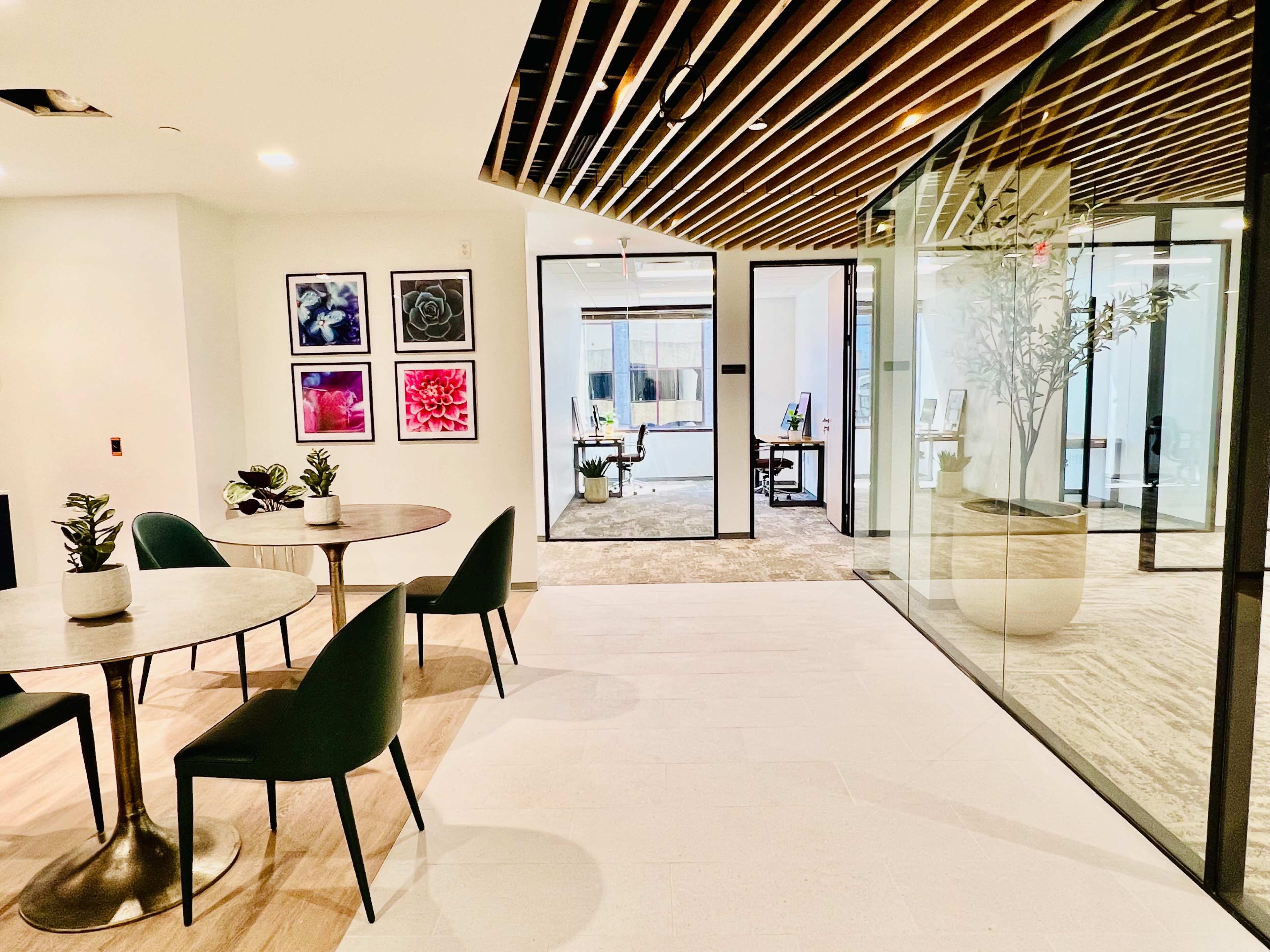 The image shows a modern office space featuring two round tables with green chairs, framed floral artwork on the walls, and a glass partition leading to another room.