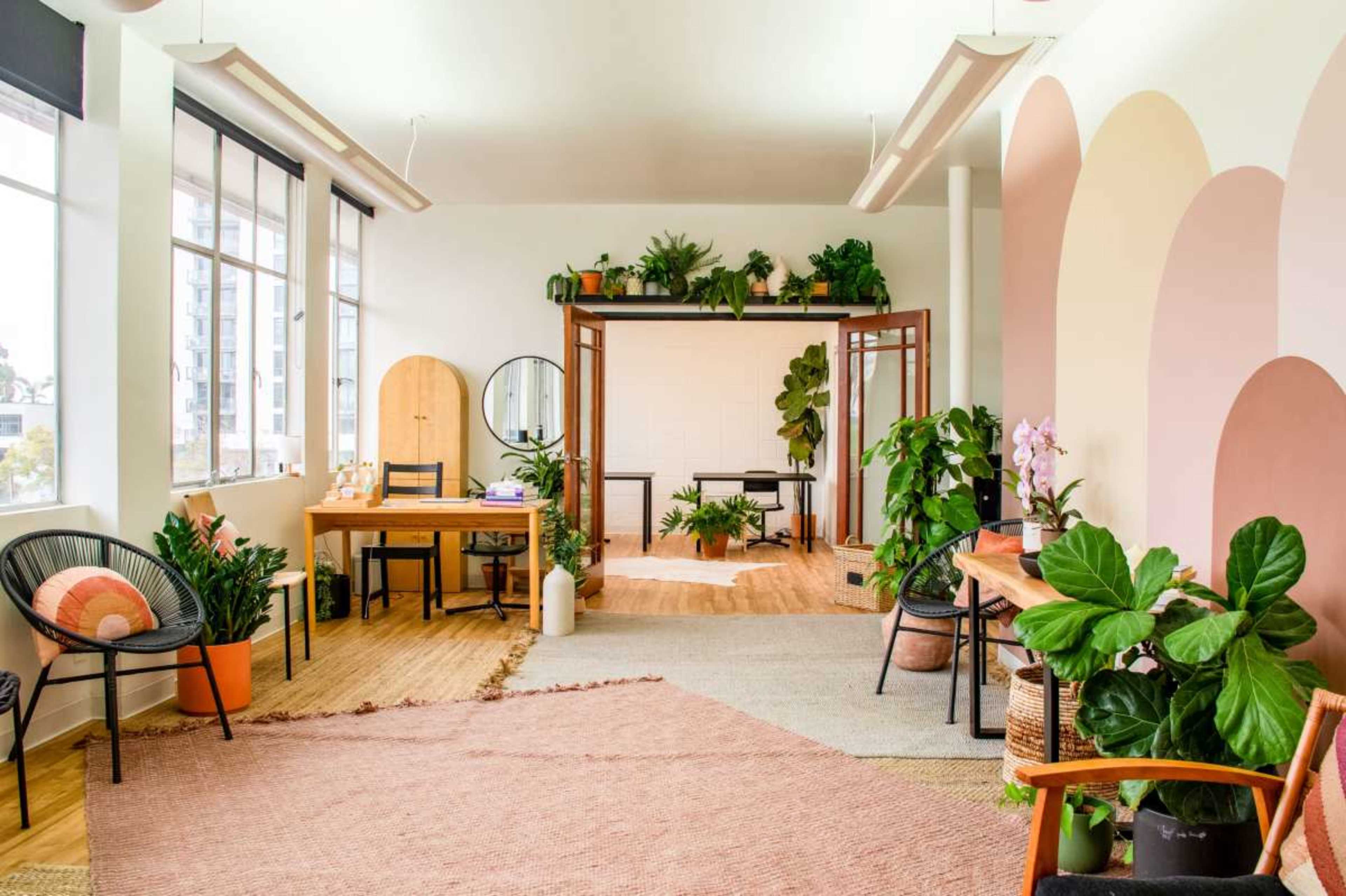 The image shows a bright, modern workspace with wooden furniture, plants, and decorative rugs arranged in an open layout.