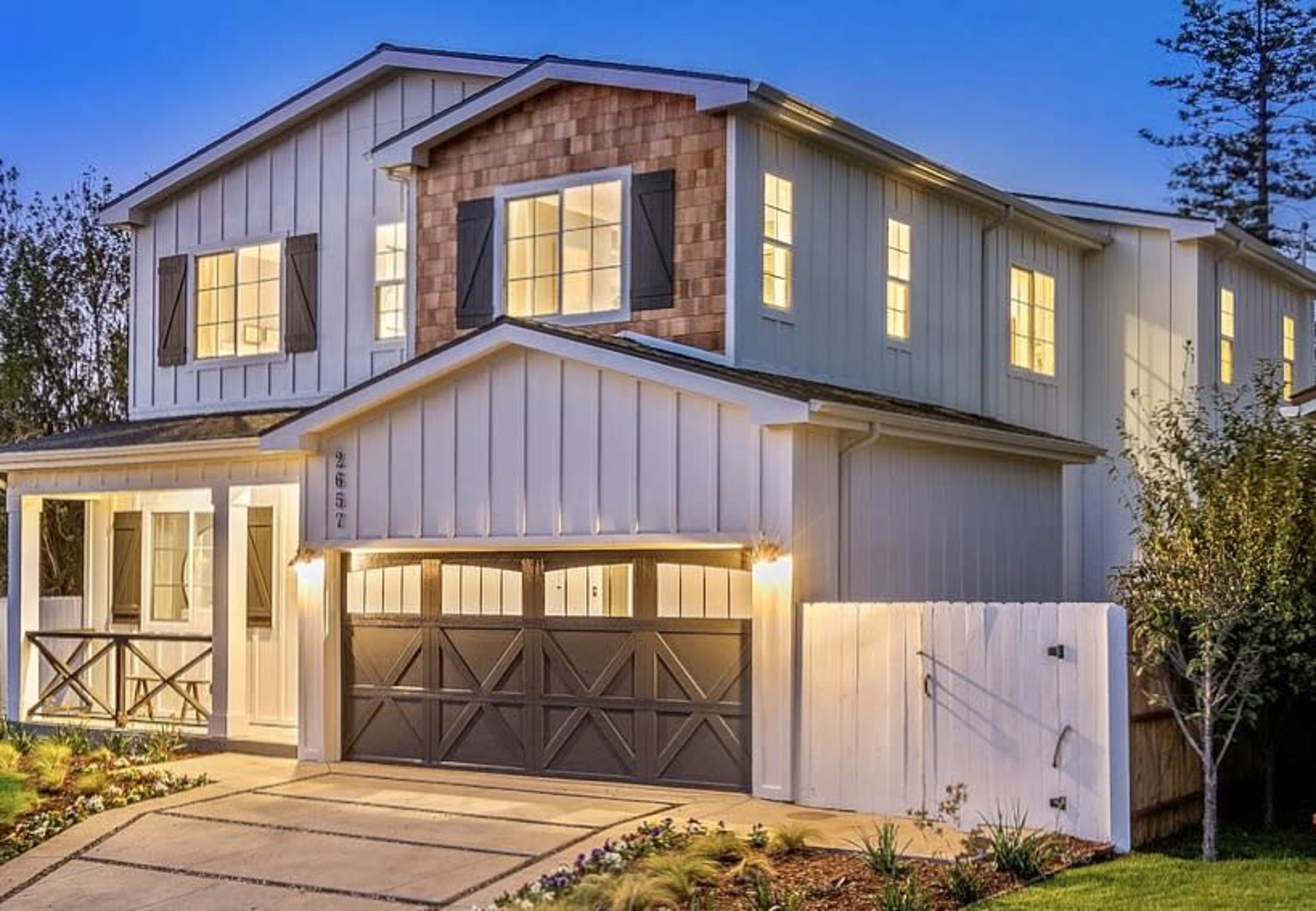 A two-story house with a combination of shingle and siding exterior features a front porch, a double garage, and landscaped greenery in the front yard, illuminated at dusk.