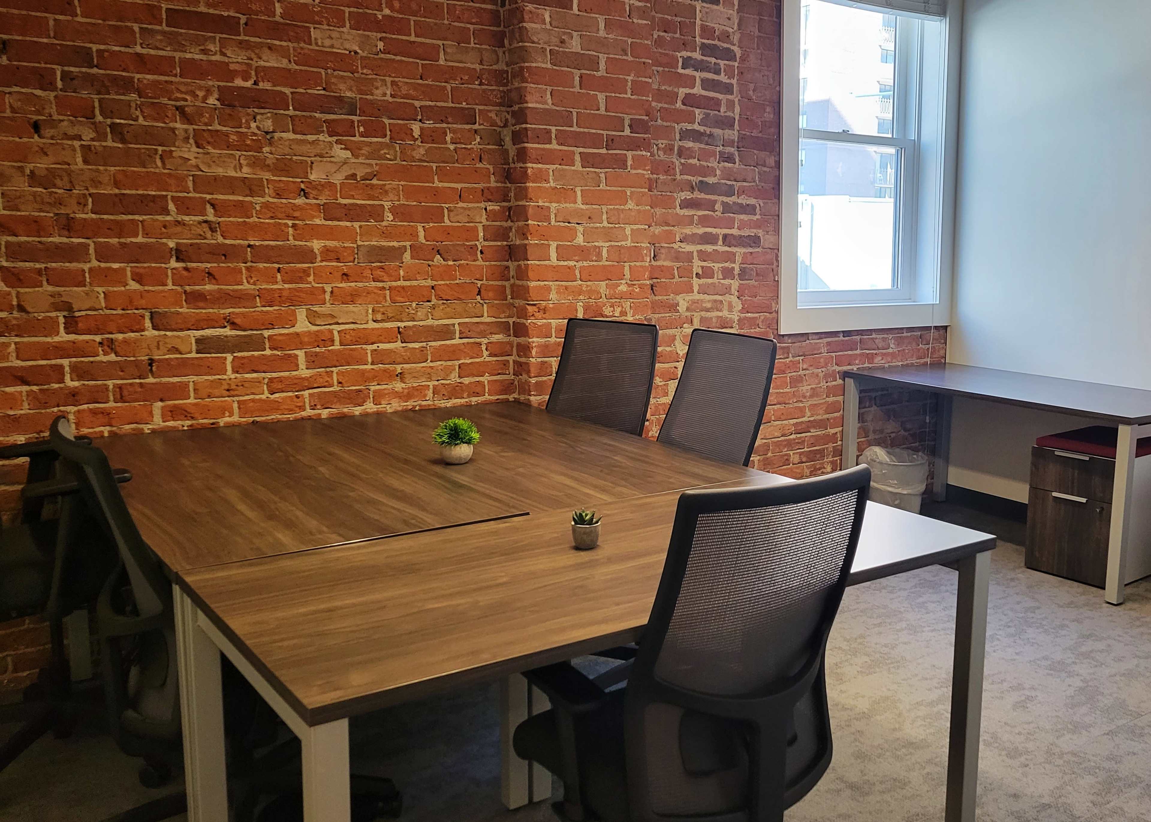 A modern office space features a wooden table surrounded by four chairs, with a brick wall in the background and a small plant on the table.