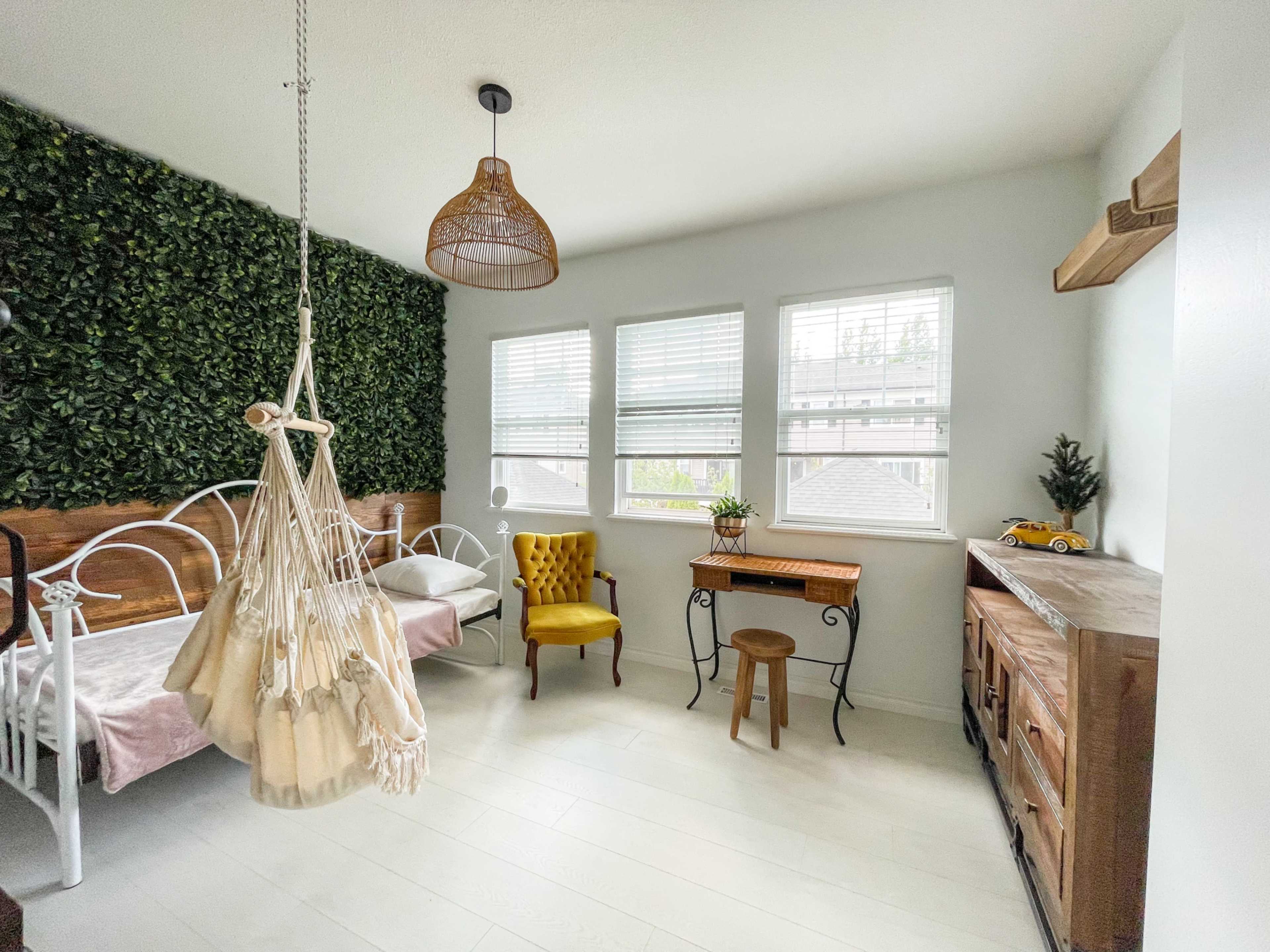 The room features two beds, a hanging chair, large windows with shades, and greenery on one wall.