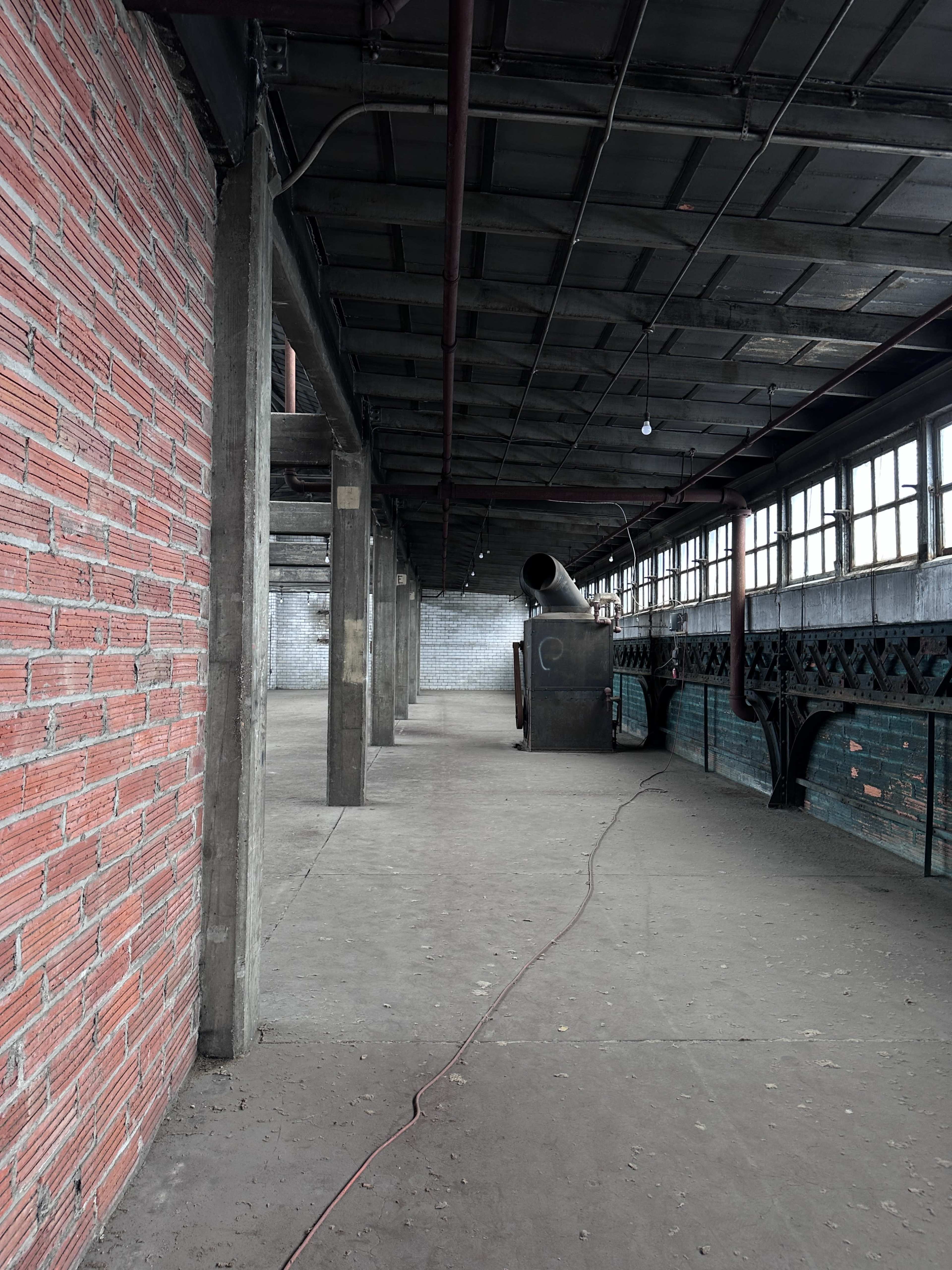 The image shows an industrial interior space with exposed brick walls, metal beams, and an empty concrete floor, featuring a large pipe extending from the wall.