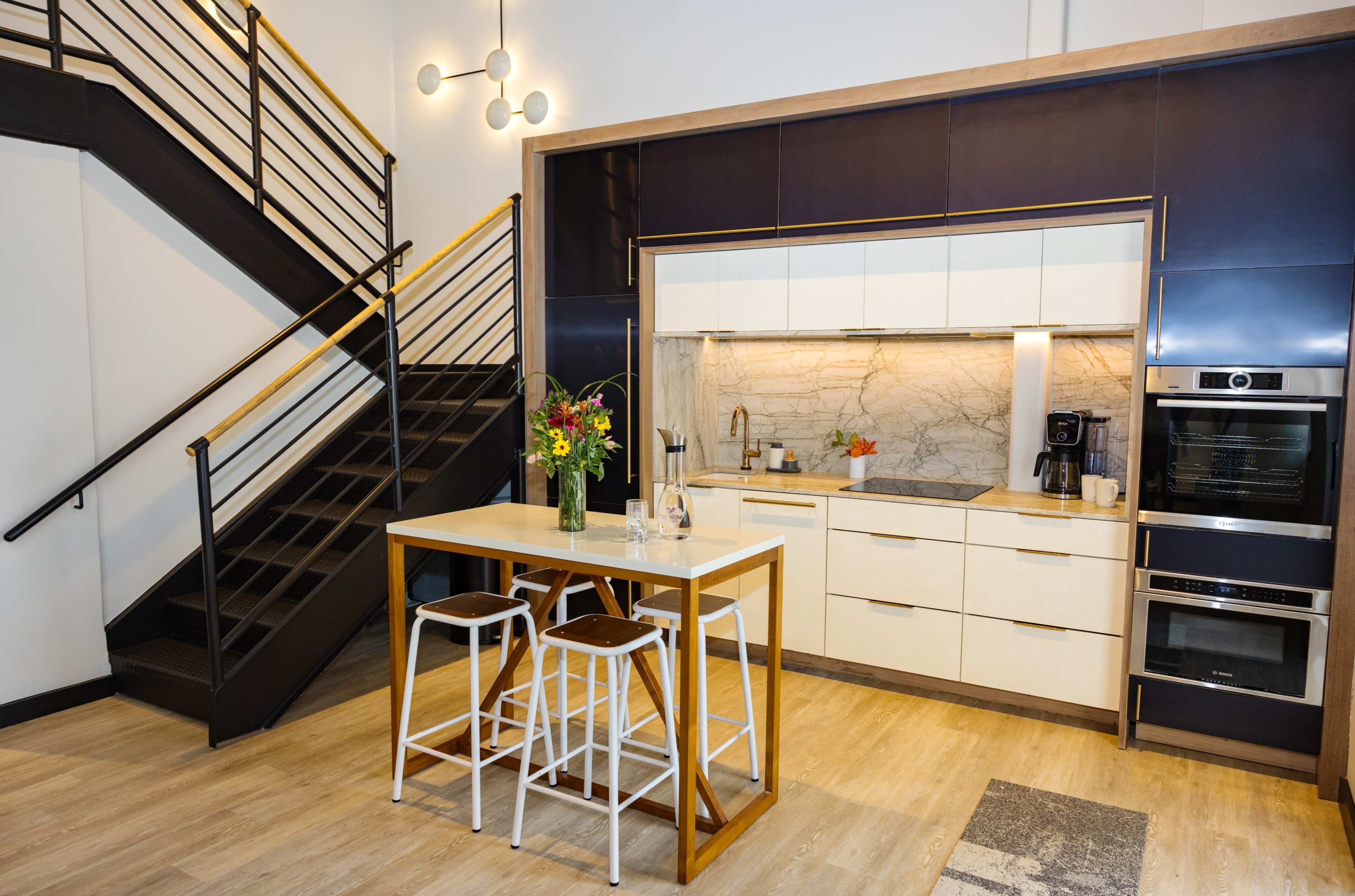 A modern kitchen features a central island with bar stools, sleek cabinetry, and a staircase in the background.