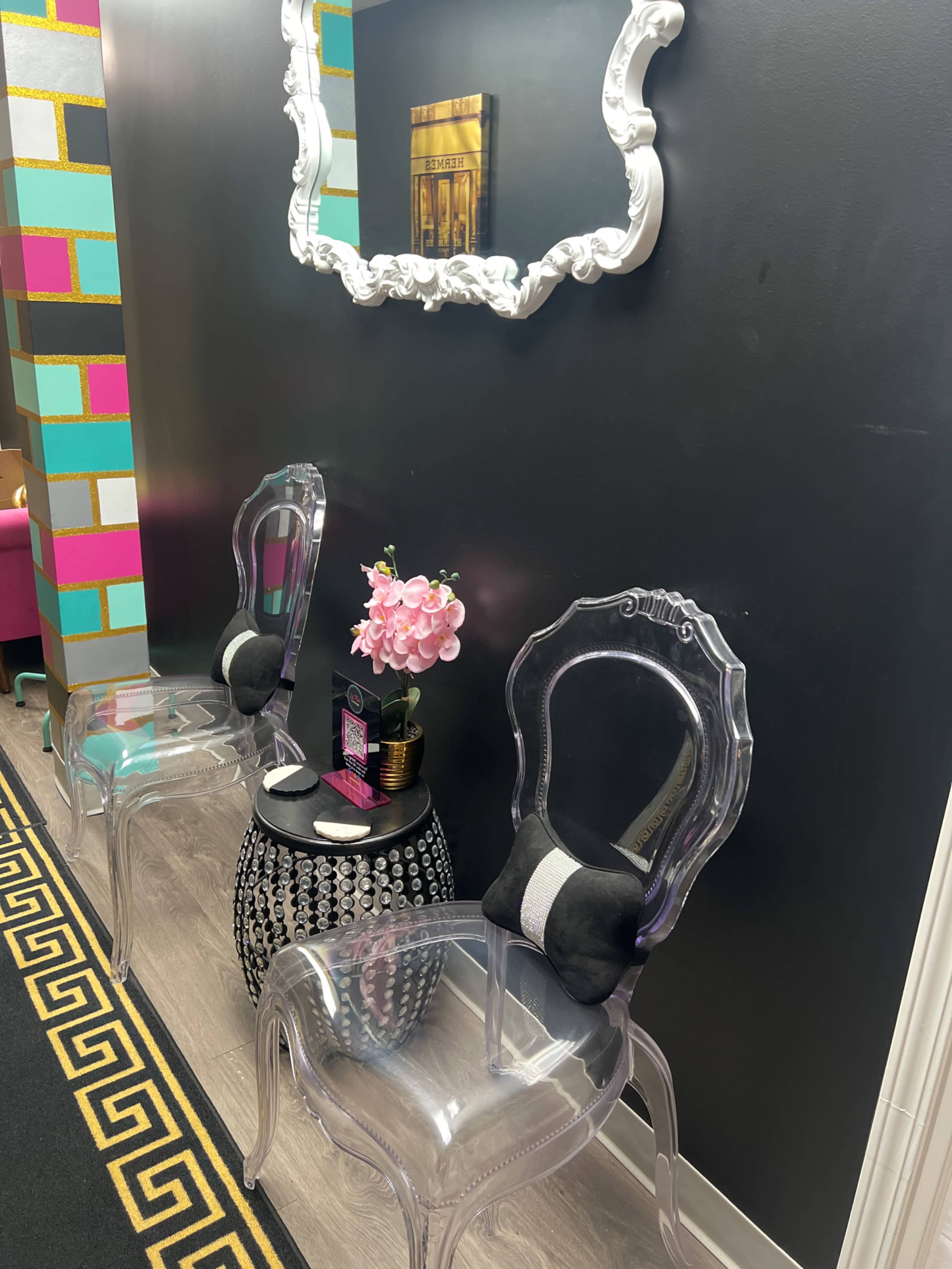 The image shows a modern waiting area featuring two clear acrylic chairs, a small round table with a decorative vase of pink flowers, and a large ornate mirror on a dark wall.