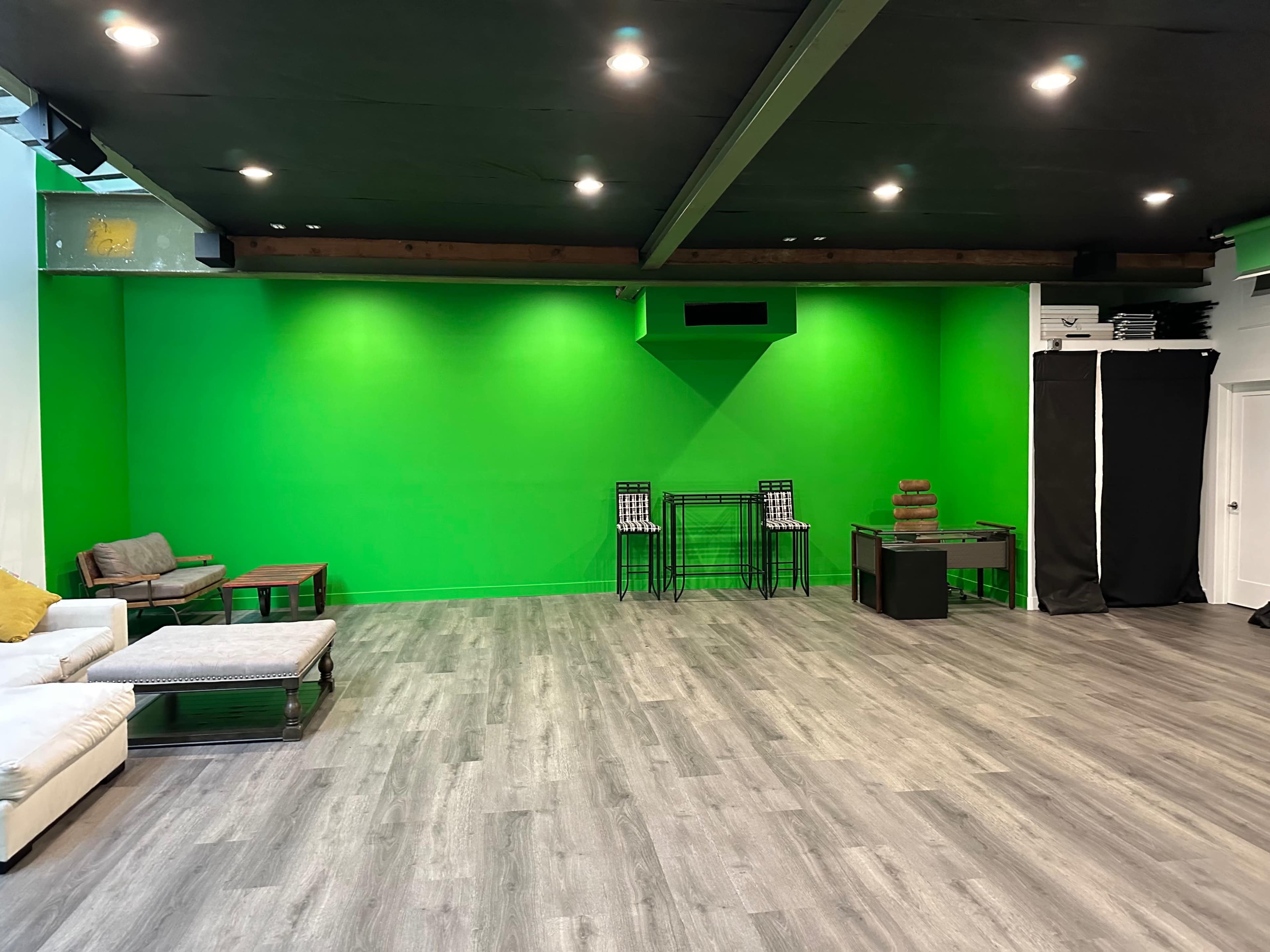 The image shows a spacious room with a large green wall, wooden beams, and a mix of seating areas and furniture.