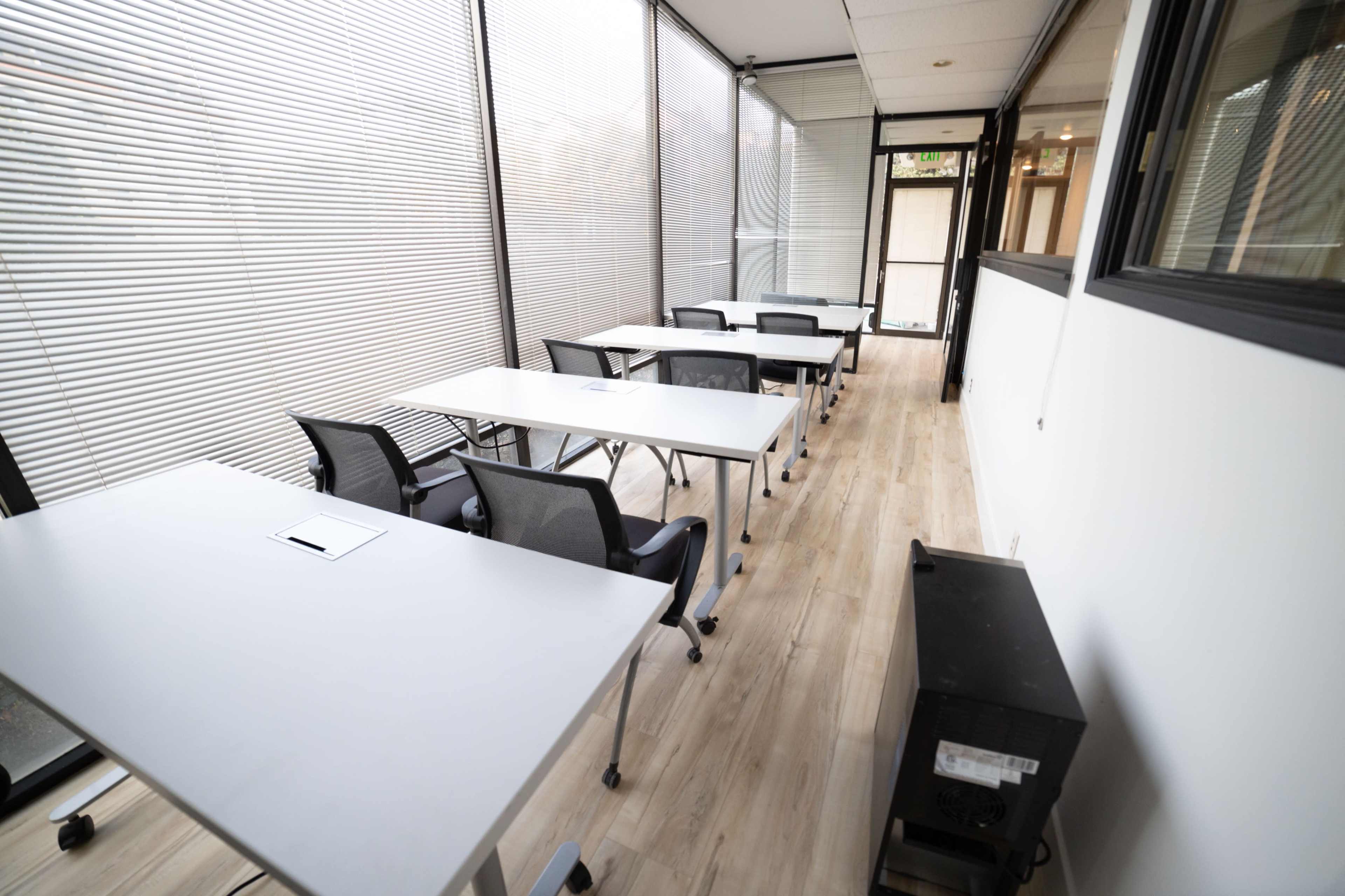 A row of tables and chairs is set up in a long, narrow classroom with large windows and light-colored wooden flooring.