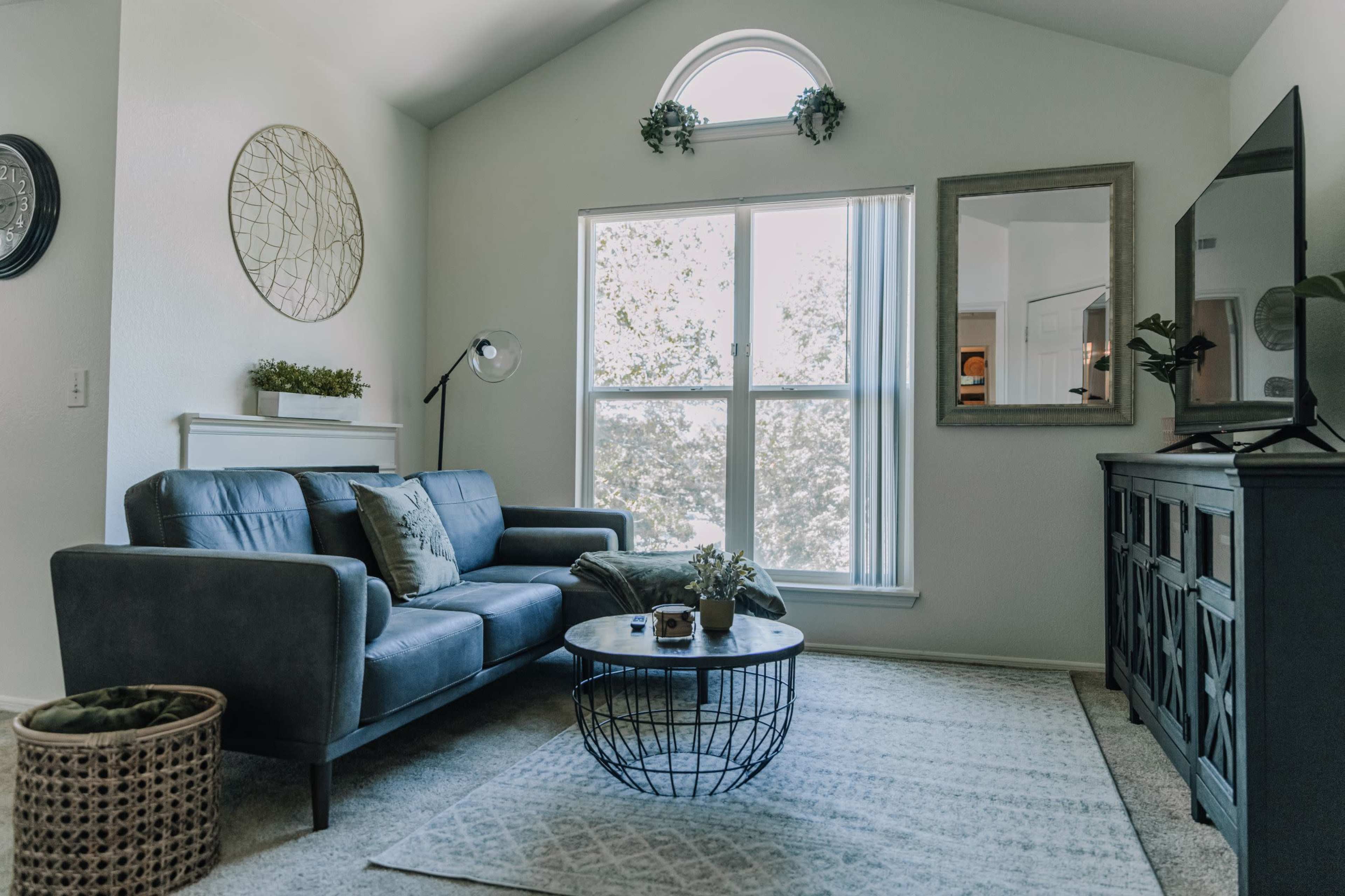A modern living room with a gray sofa, a round coffee table, a decorative rug, and a large window allowing natural light to enter.