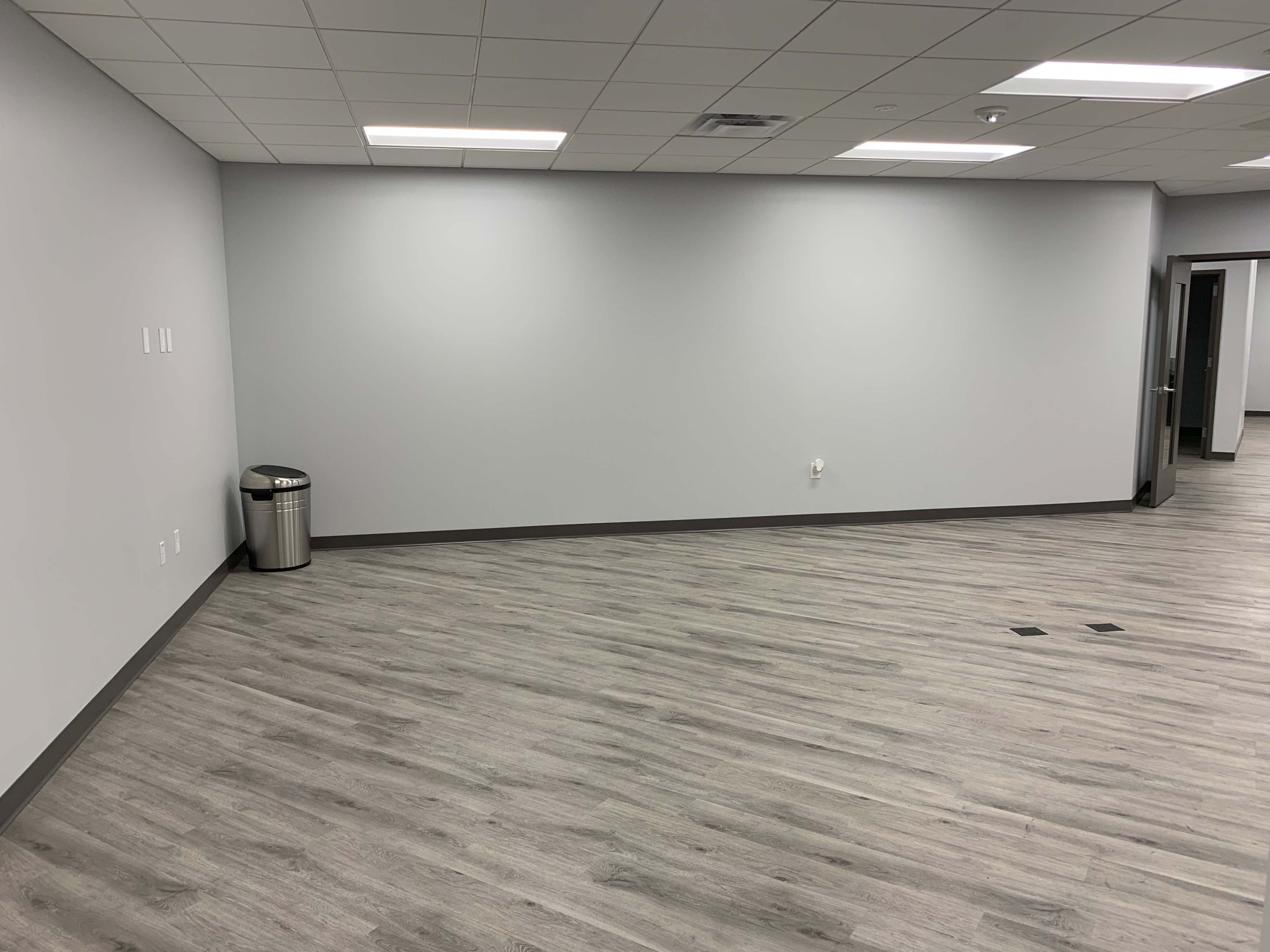 The image shows a vacant office space with gray walls, a wooden floor, a trash can in the corner, and a doorway on the right.