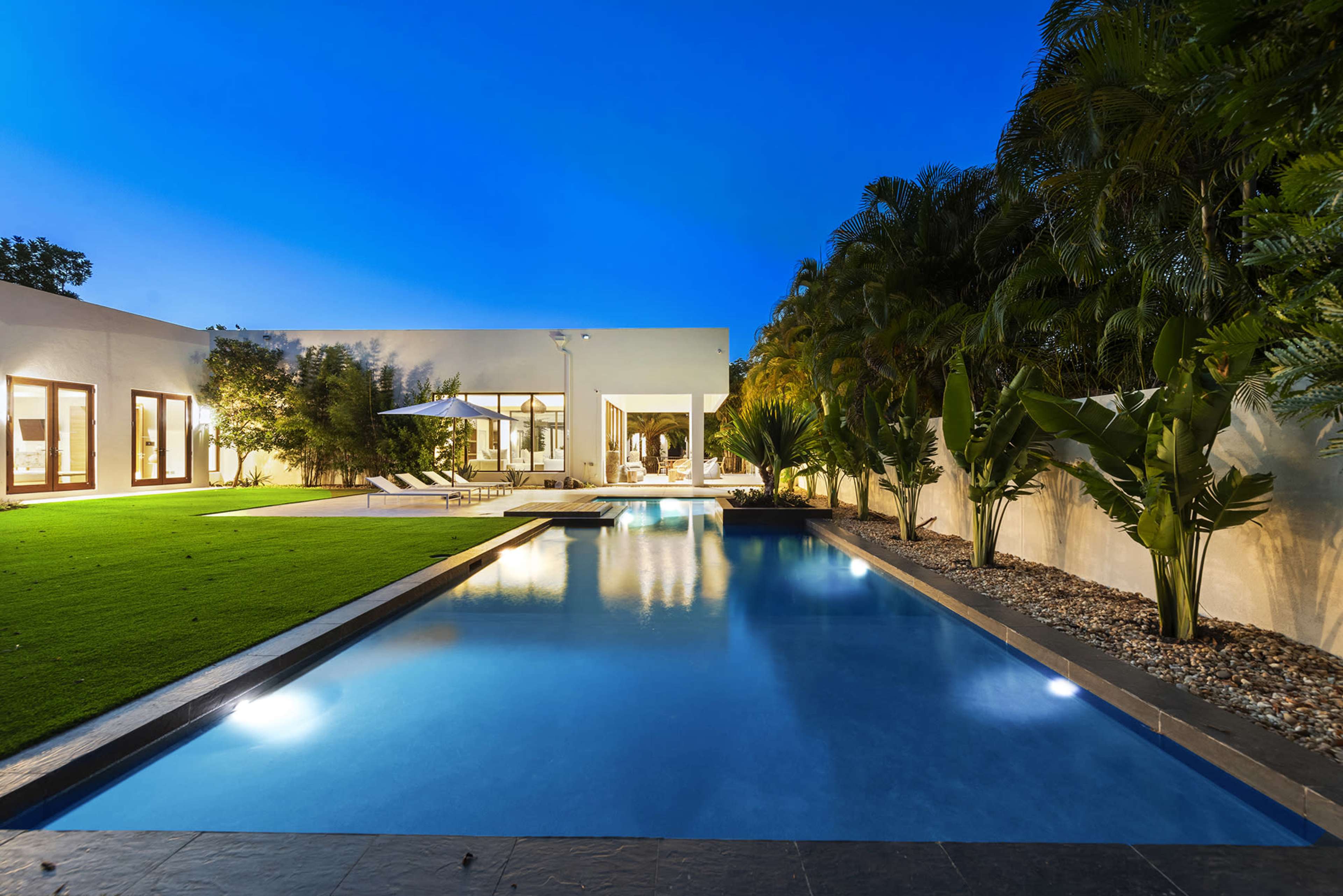 The image displays a modern home with a swimming pool, green lawn, and tropical plants under a twilight sky.