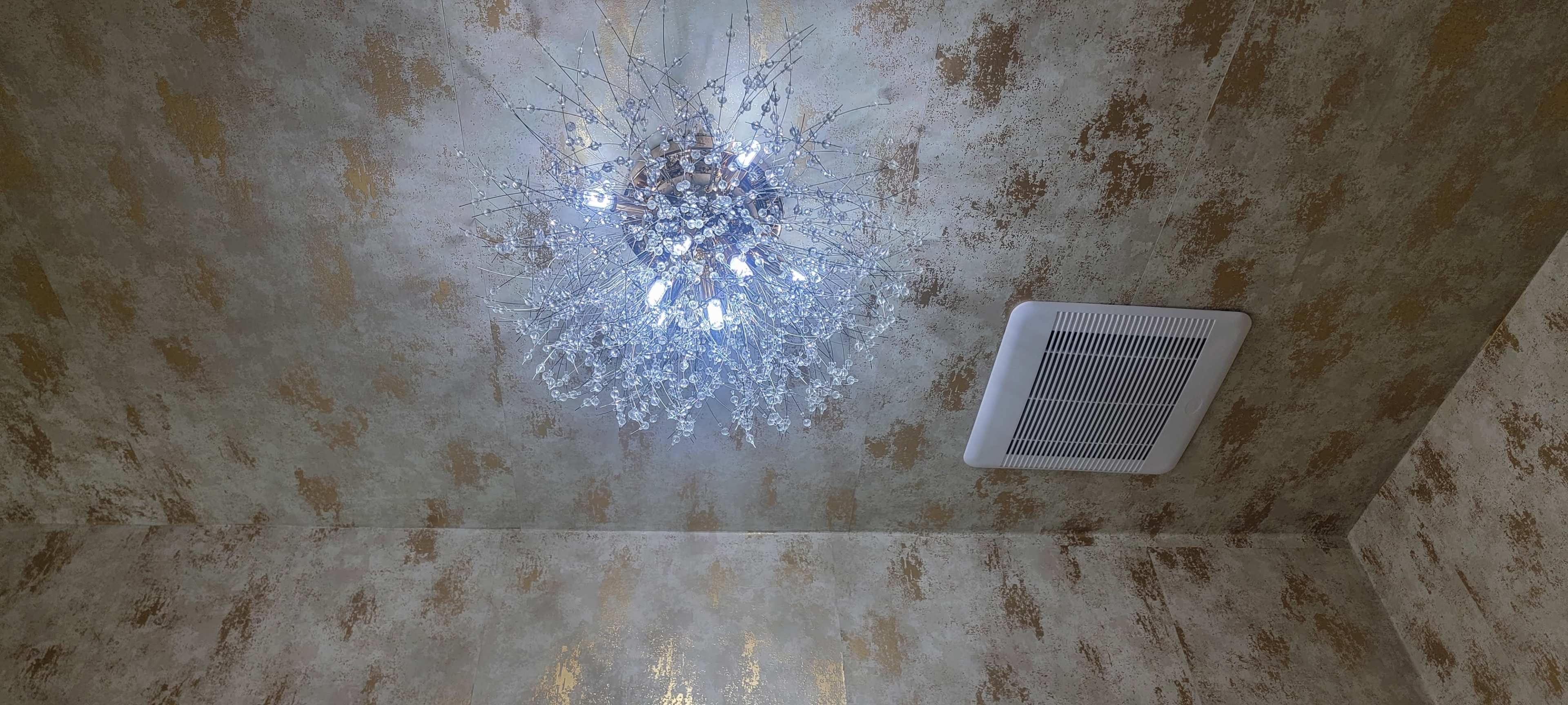 A modern chandelier hangs from a textured ceiling with a gold and cream patterned finish, alongside a ventilation grill.