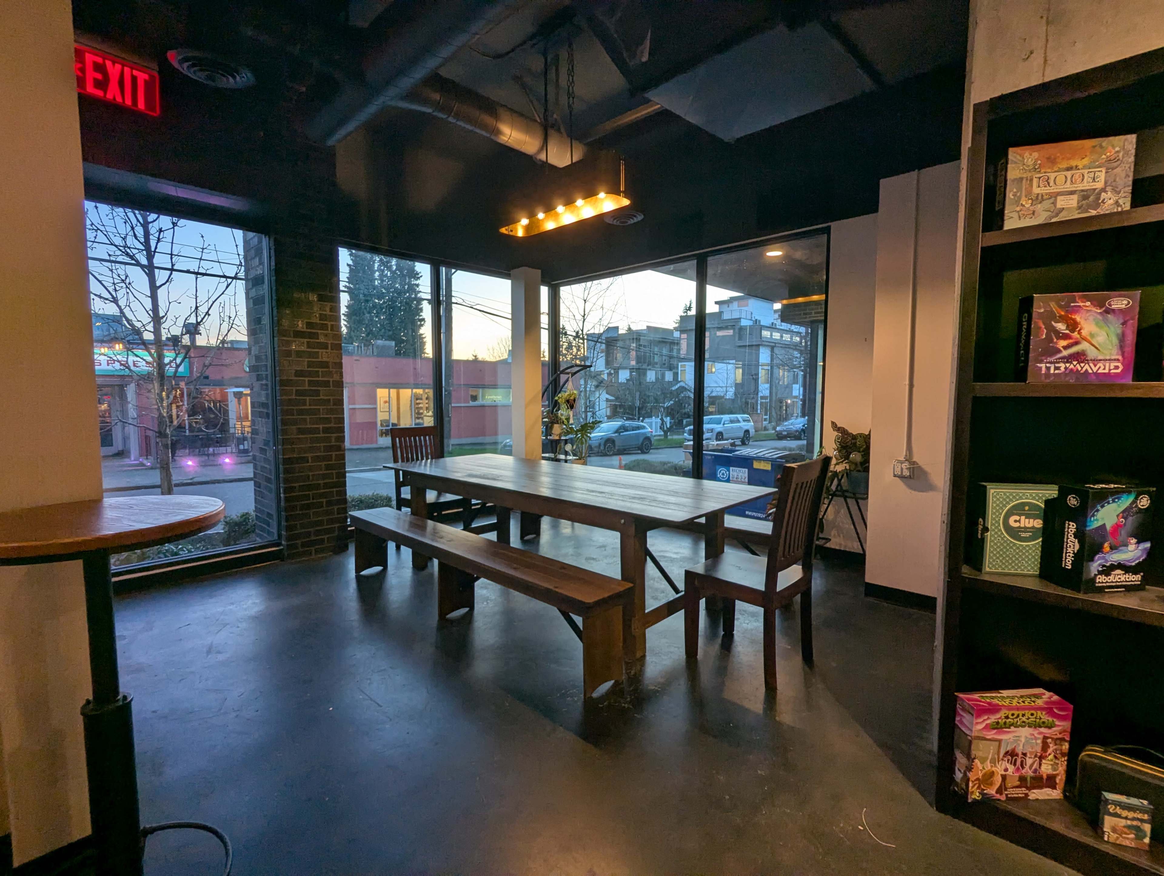 The scene shows a spacious room with large windows, a long wooden table, and chairs, offering a view of a street outside during dusk.