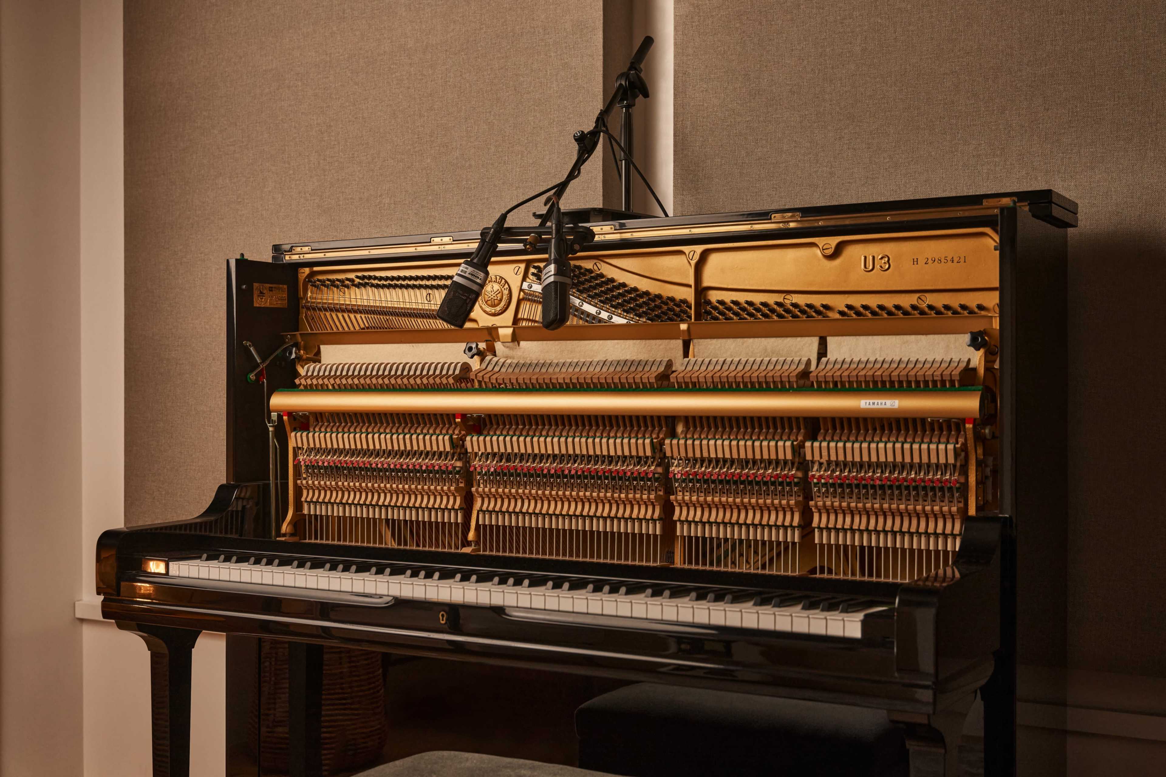 A black upright piano is placed against a wall, featuring exposed strings and two microphones positioned above the soundboard.