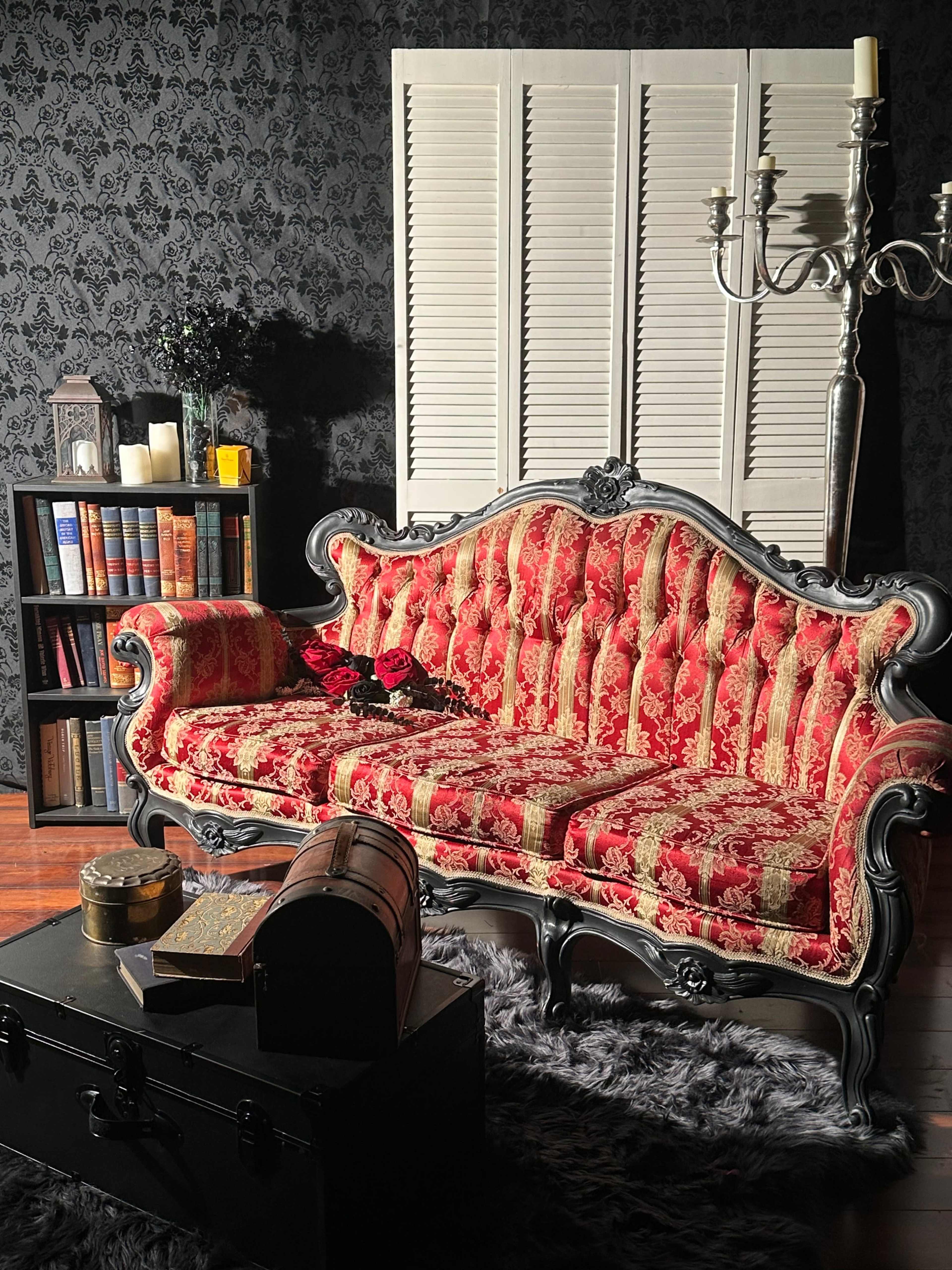 The image features an ornately designed red and gold patterned Victorian-style sofa, positioned near a bookshelf filled with books and decorative items, under soft lighting that creates a cozy atmosphere.