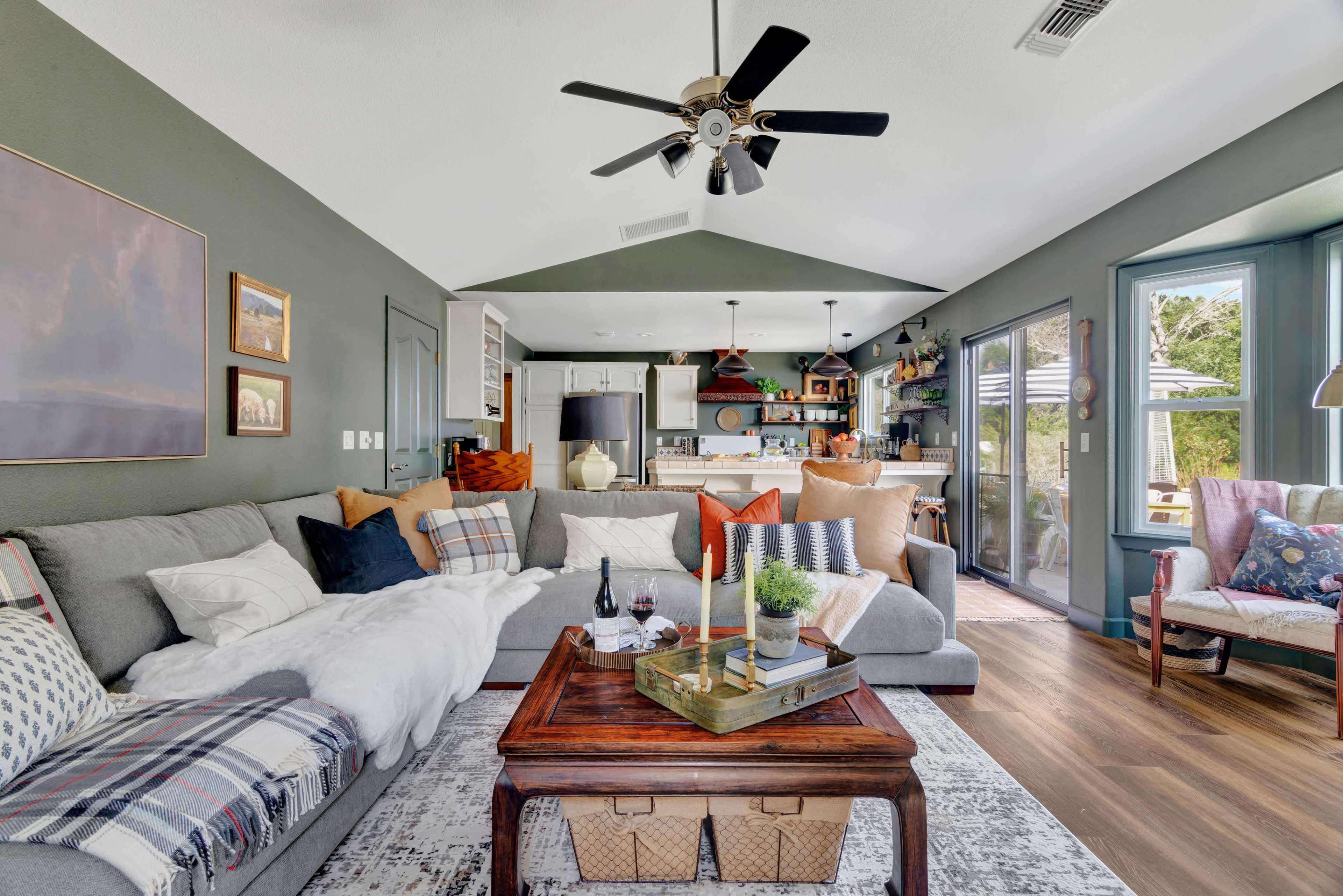 A cozy living room features a large gray sectional sofa, a wooden coffee table, and open access to a bright kitchen area.