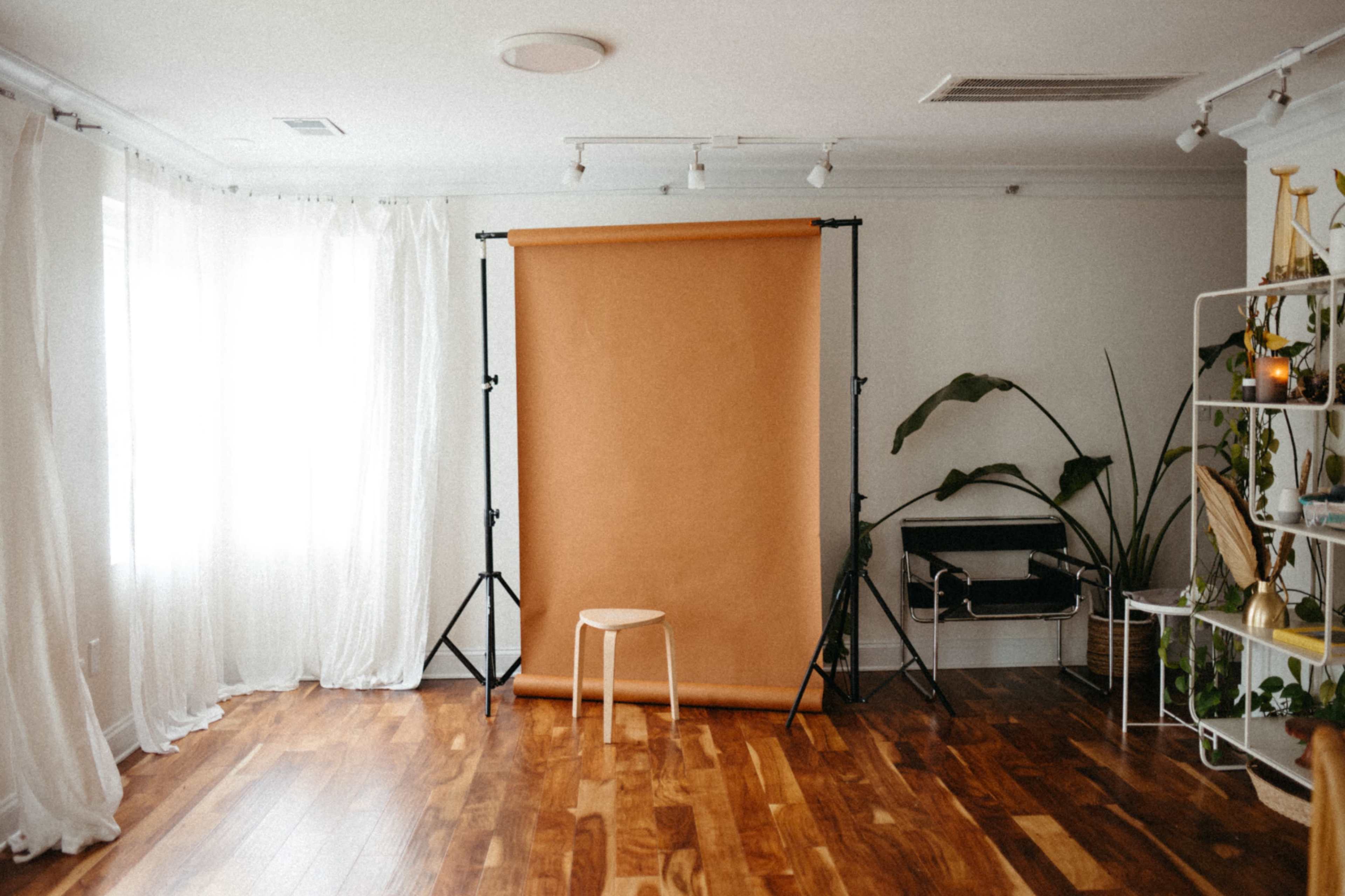 The image shows a photography studio with a brown backdrop on a stand, a wooden stool in front of it, and potted plants and furniture in the background.