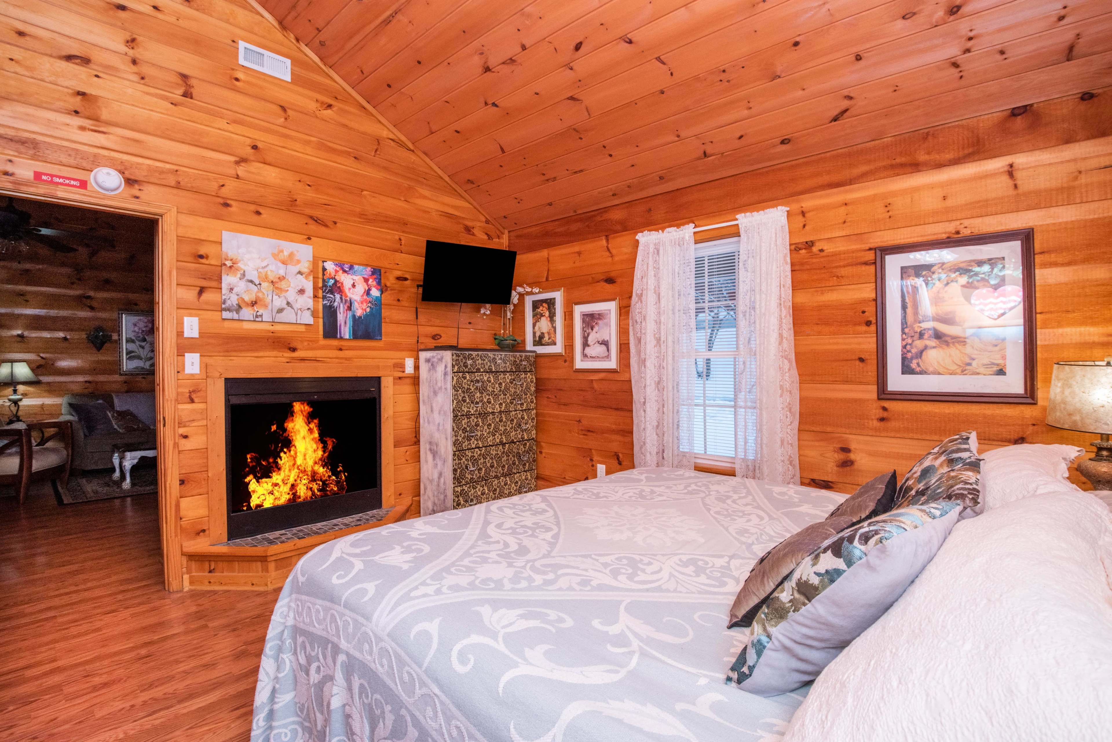 A cozy bedroom with wooden walls, a bed with decorative pillows, a television mounted on the wall, and a fireplace with flames.
