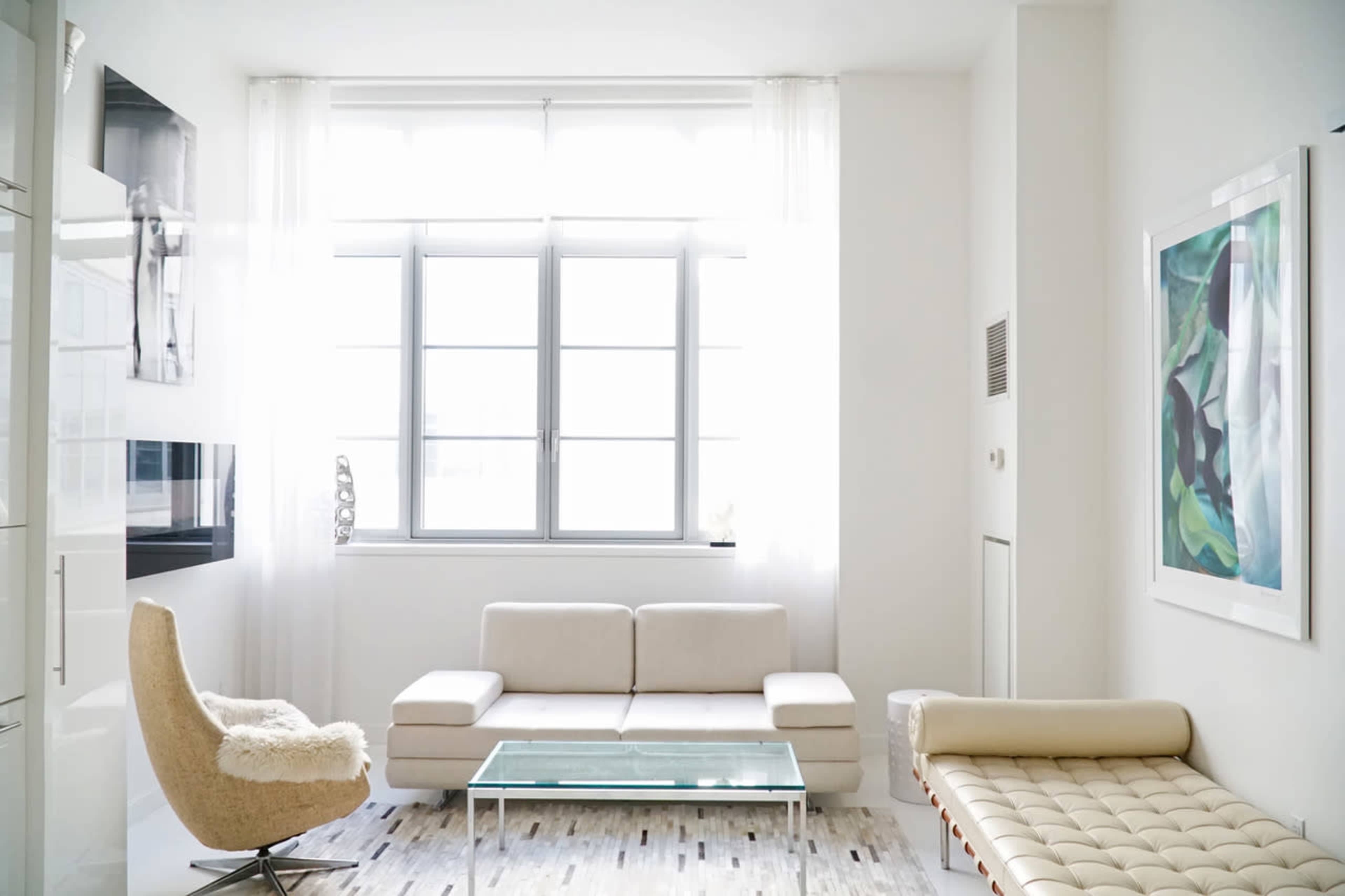A bright, modern living room featuring a light-colored sofa, a glass coffee table, and large windows with sheer curtains.