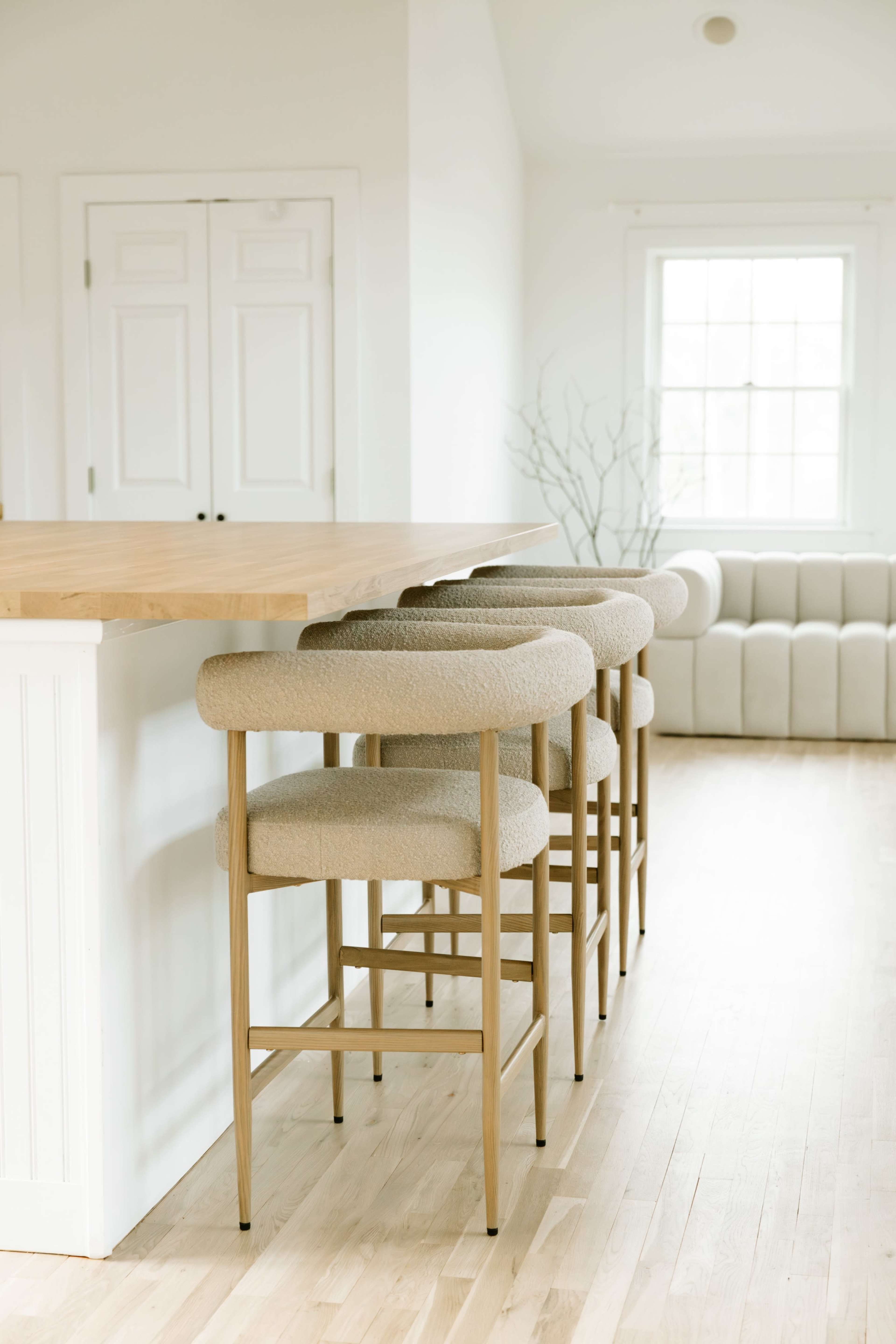A wooden kitchen island with four upholstered bar stools is positioned near a white wall and a light-colored sofa in a bright, airy room.