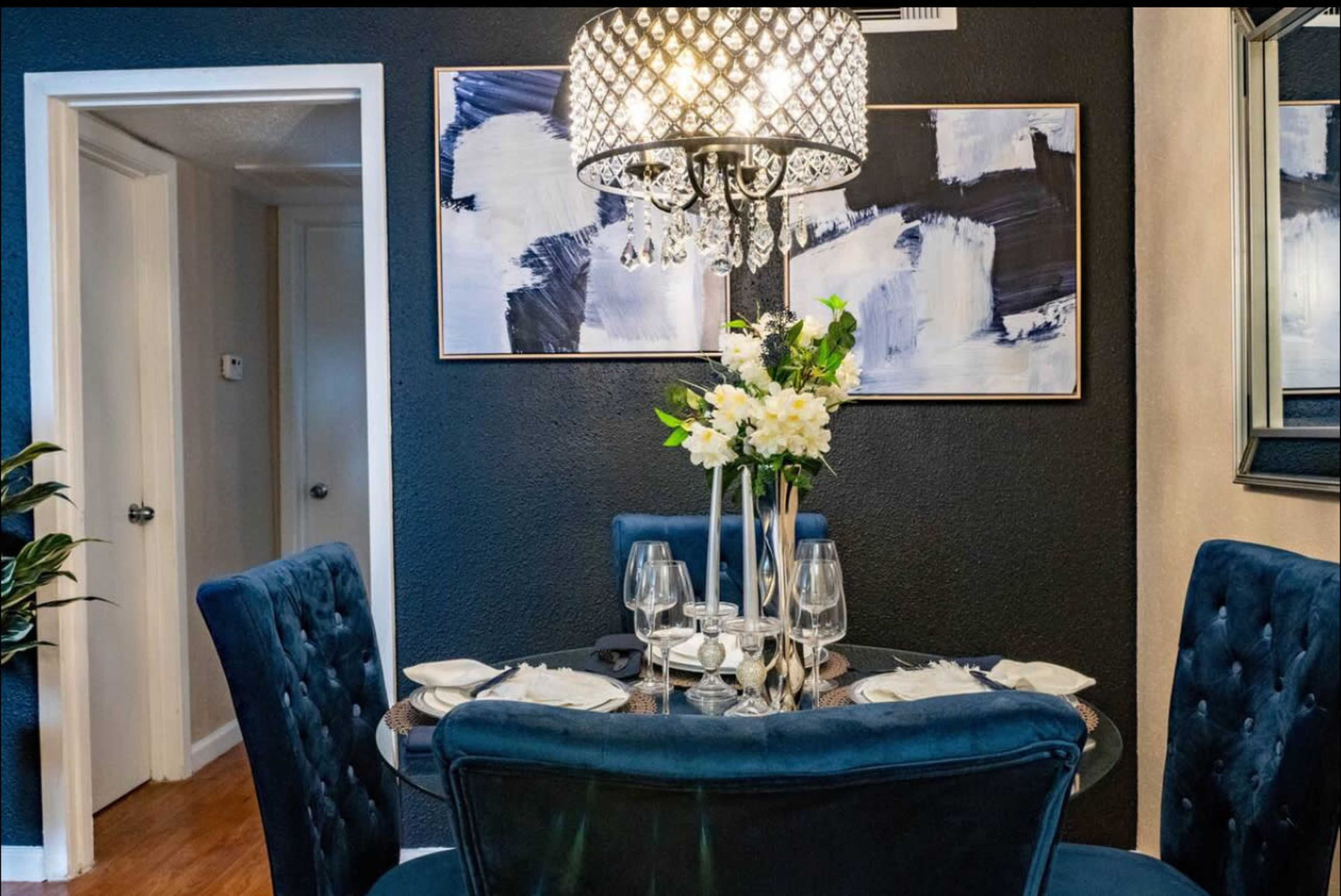 A round dining table is set with plates and glasses, surrounded by upholstered chairs, beneath a decorative chandelier in a stylishly decorated room.