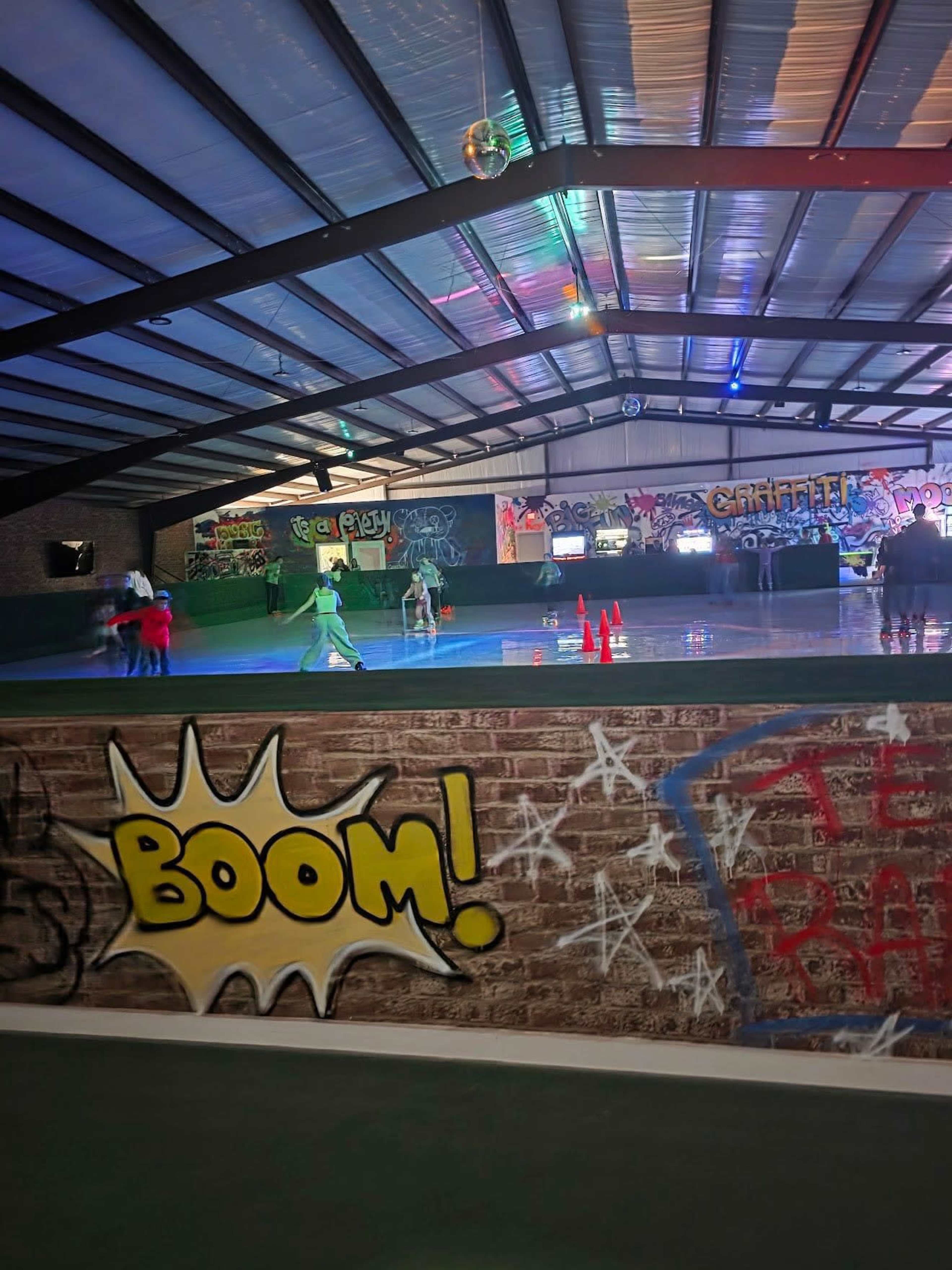 The image shows an indoor skating rink with colorful graffiti on the walls and several people skating on the rink.