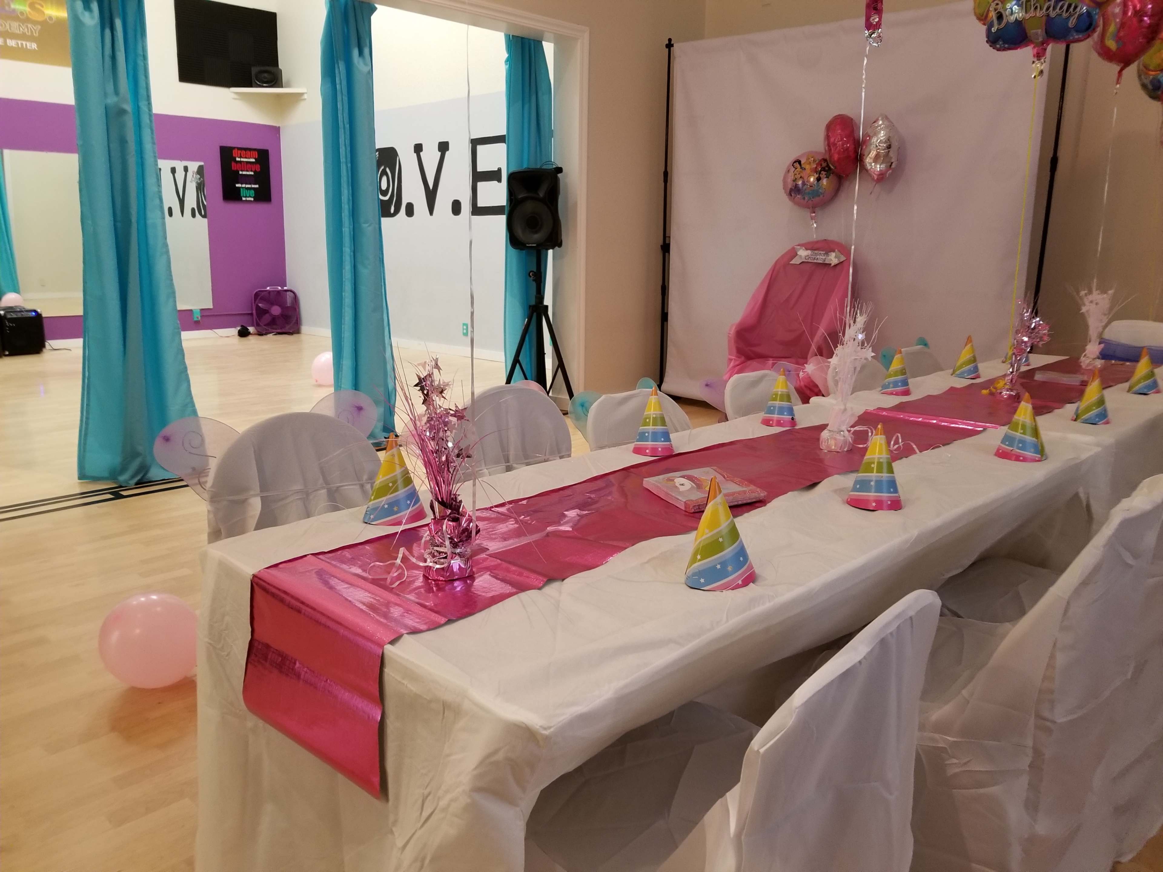 A decorated party room with a long white table set with colorful party hats and a pink centerpiece, surrounded by white chairs and balloons.