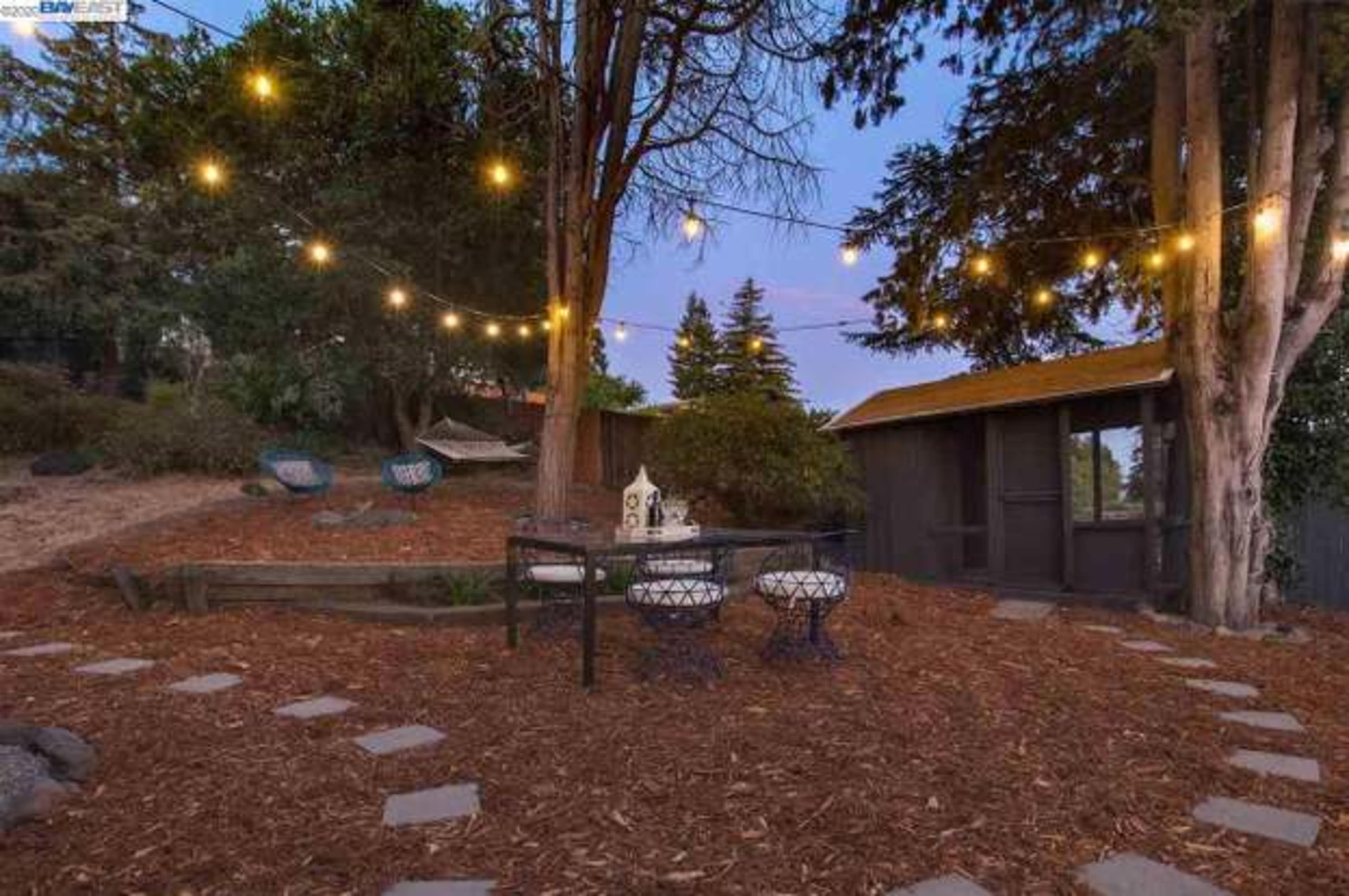 A backyard patio area with string lights overhead, a table and chairs, and decorative gravel paths surrounded by trees and shrubs.