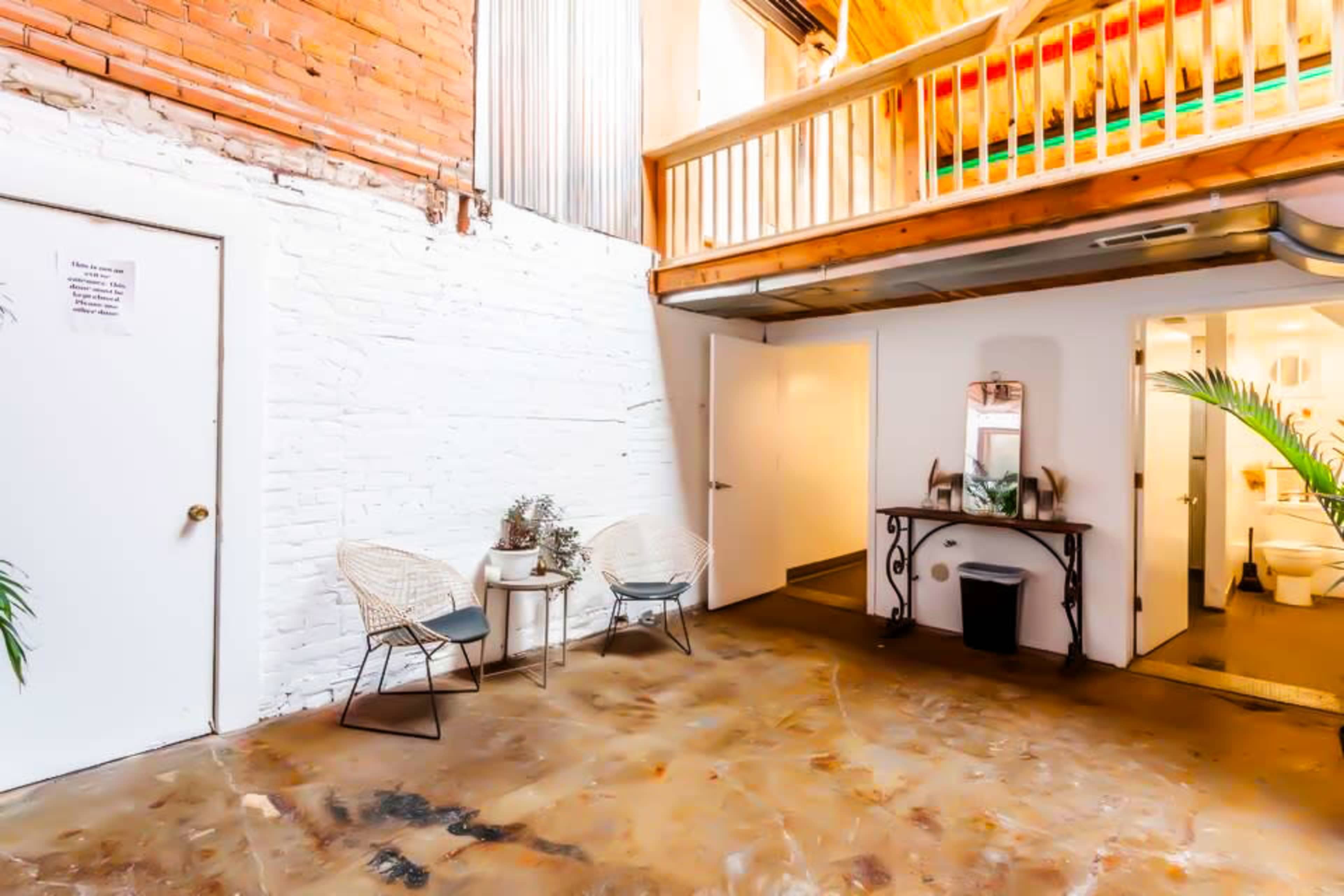 The image shows a spacious interior with exposed brick walls, a seating area consisting of two chairs and a table, and access to a bathroom in a well-lit setting.