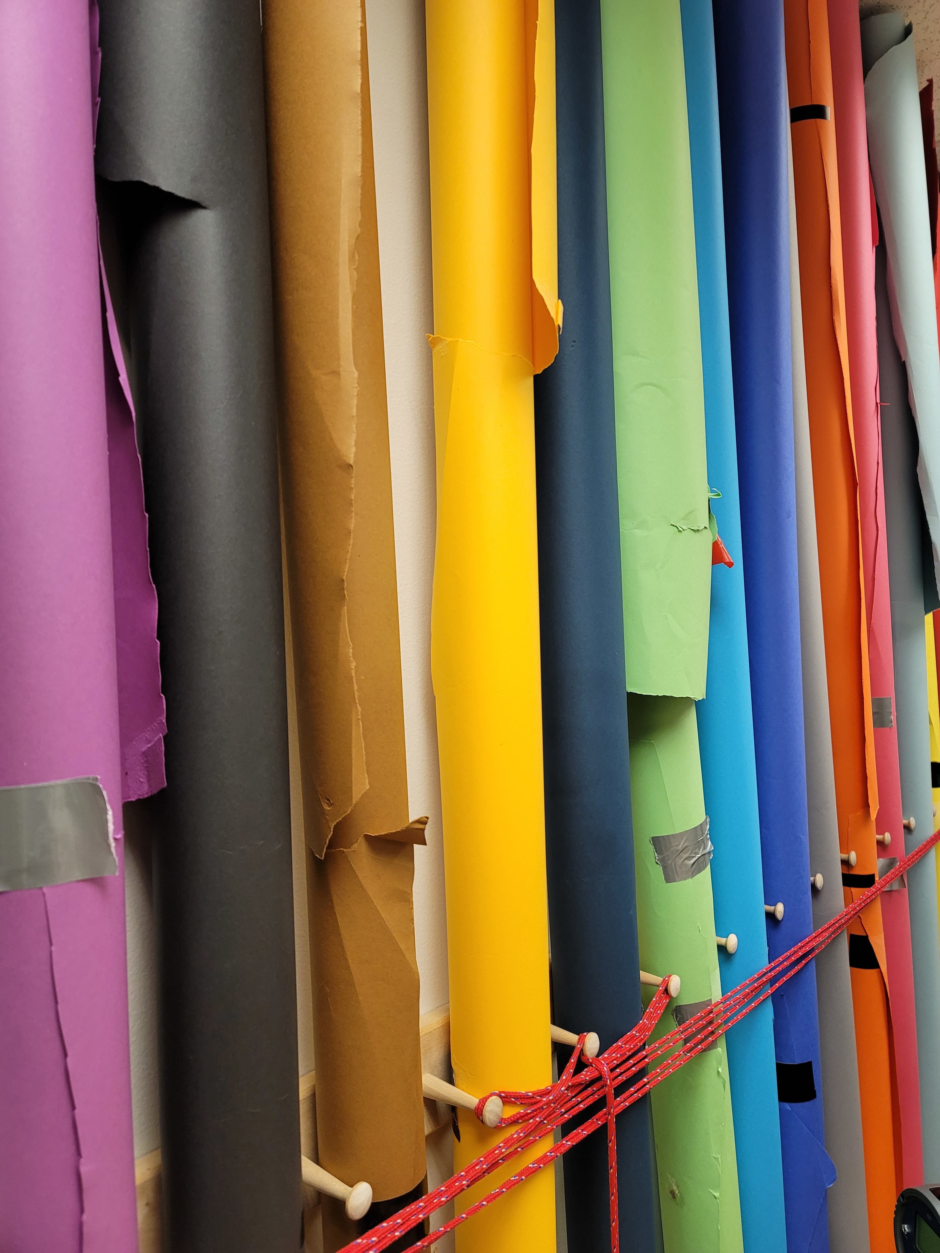 A collection of colorful rolls of paper is displayed on a wall, held in place by wooden dowels and tied with red string.