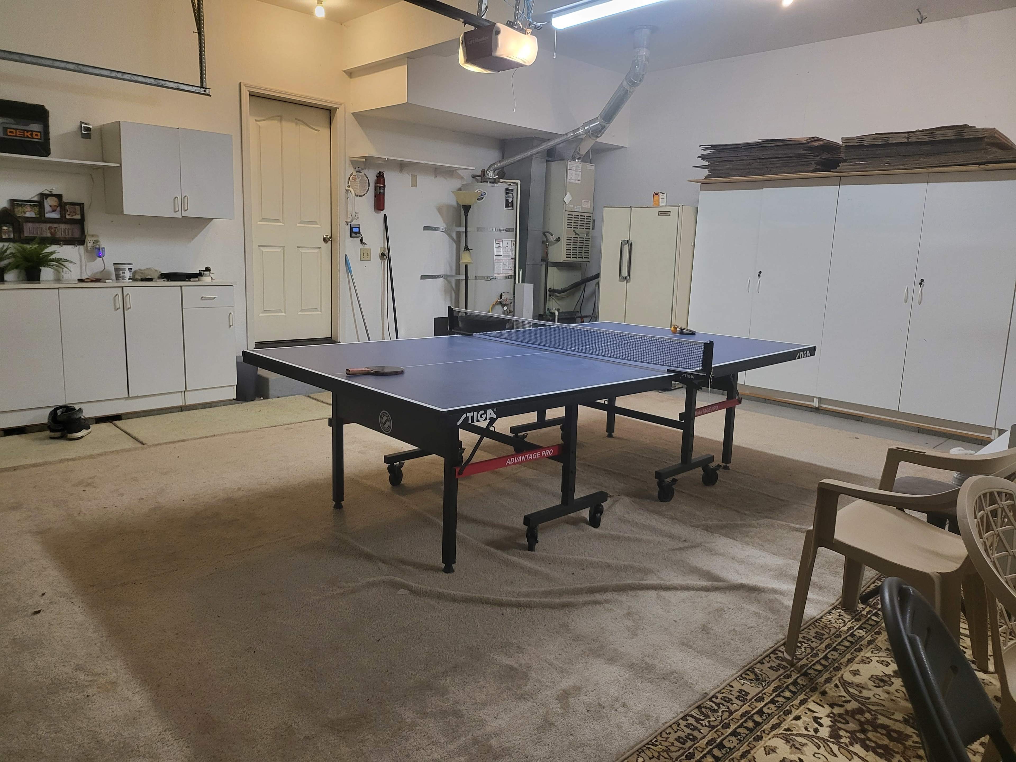 A ping pong table is set up in a garage, surrounded by chairs, cabinets, and tools.