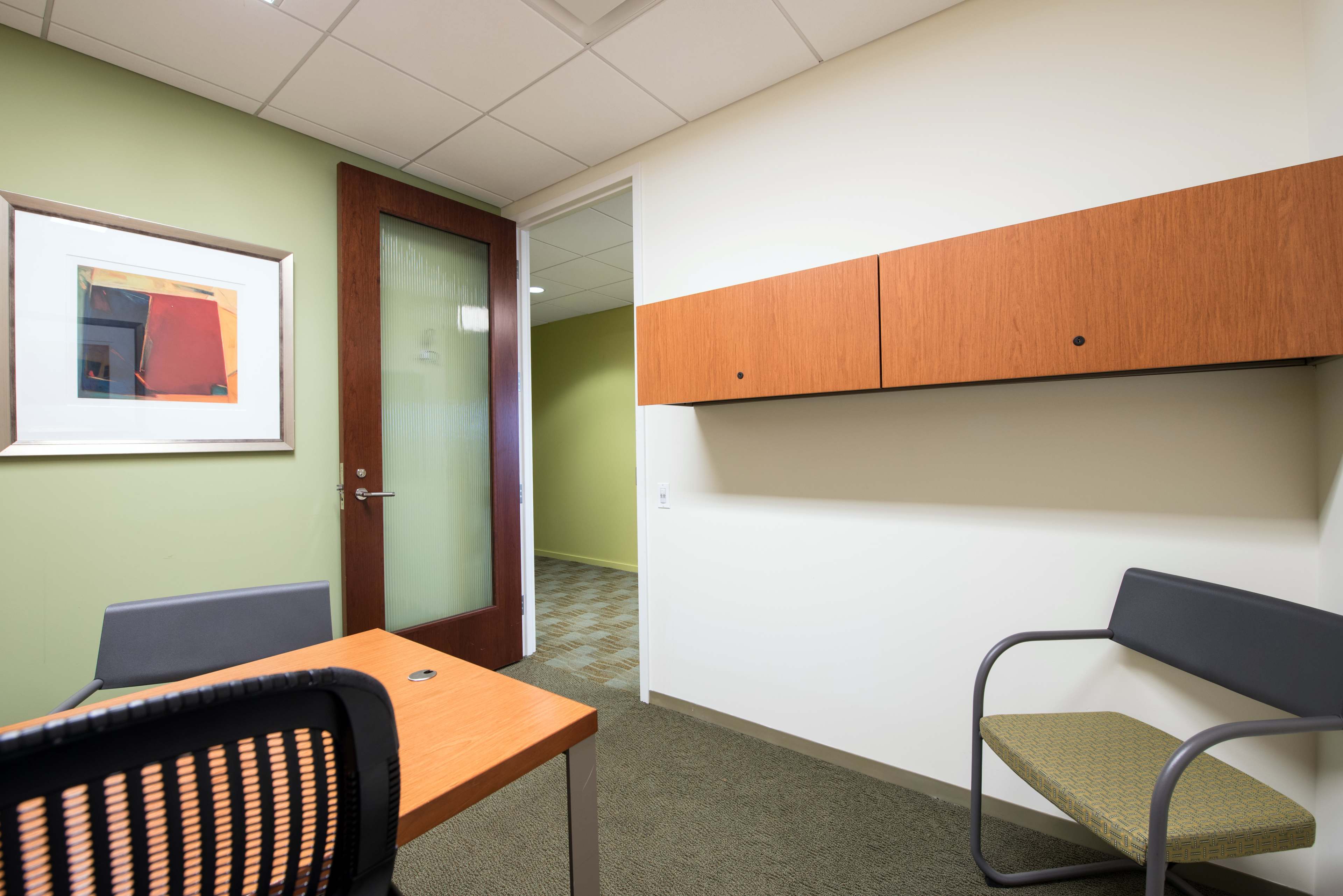 A small office space featuring a wooden table, a chair, a wall-mounted storage unit, and a glass-paneled door leading to another room.