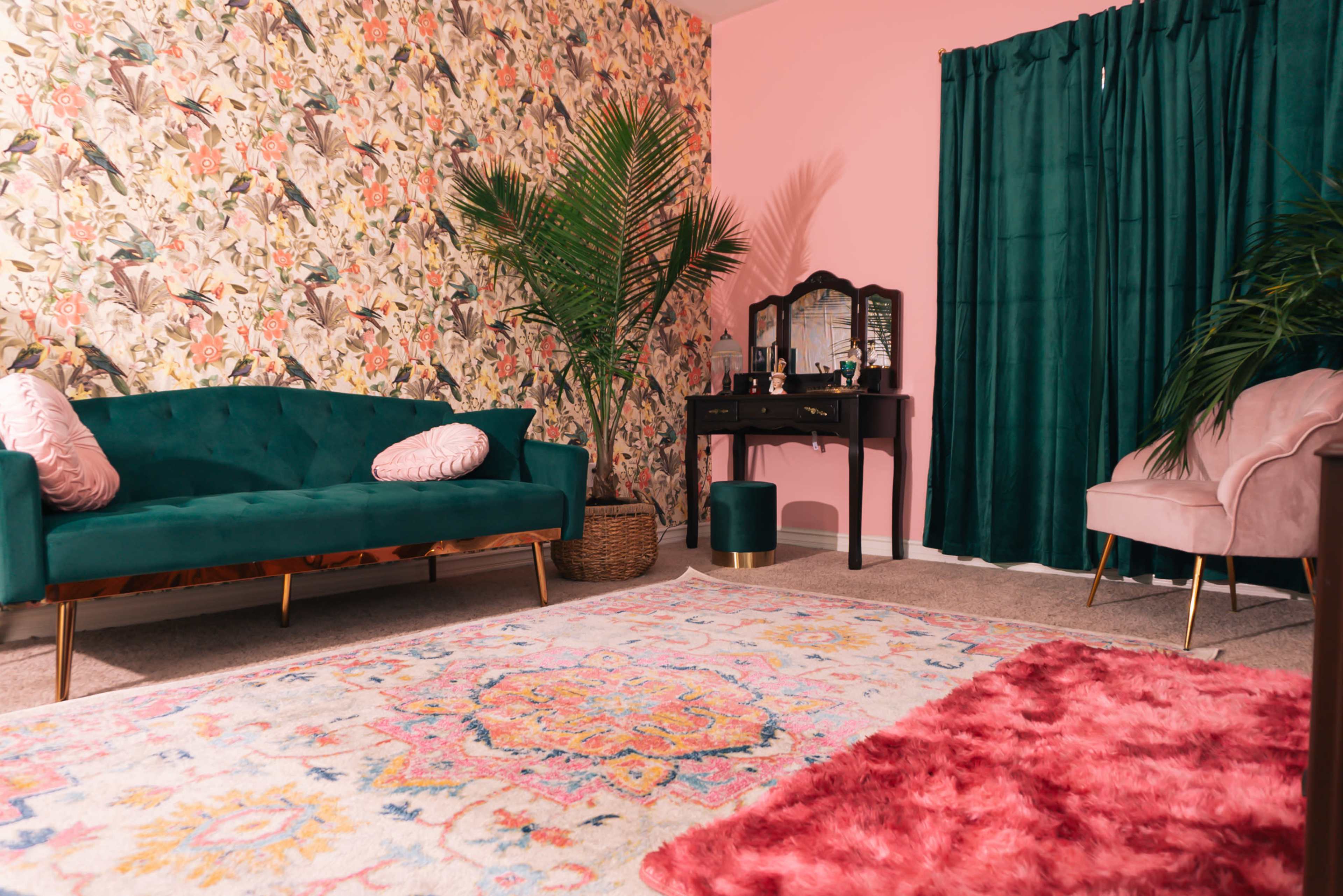 A colorful living room with a patterned wallpaper, a green sofa, a black vanity table, and a pink chair, complemented by a large area rug and plants.