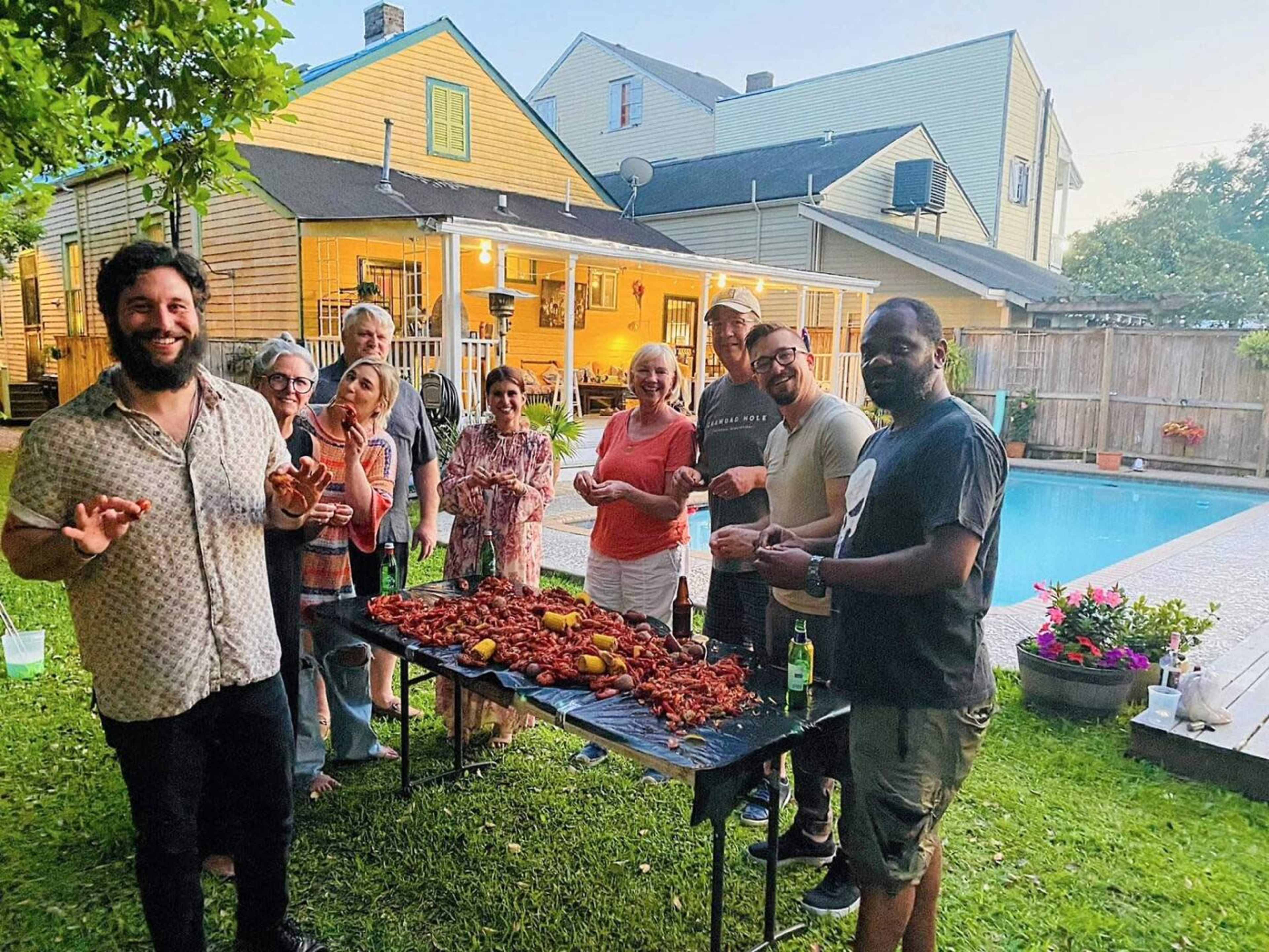 A group of people stands around a table filled with seafood in a backyard with a pool and houses in the background.
