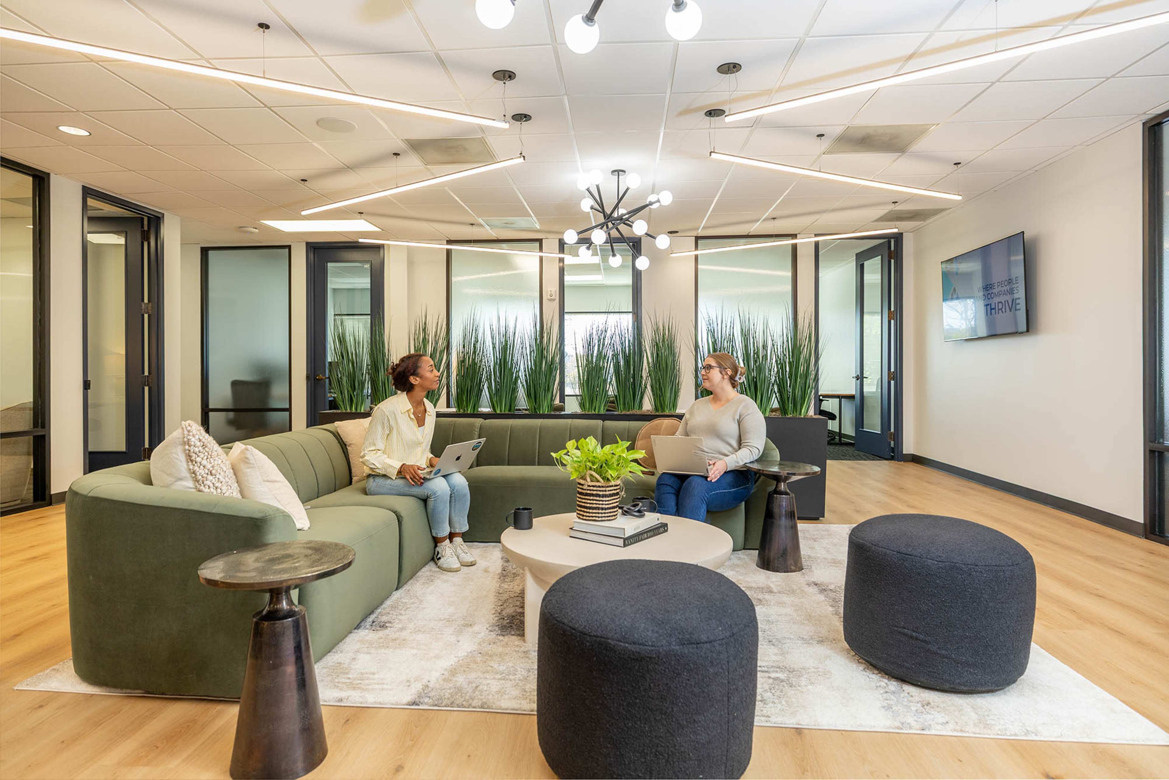 Two women are seated in a modern lounge area with a green sofa, round coffee table, and decorative plants, engaging in conversation while using a laptop.