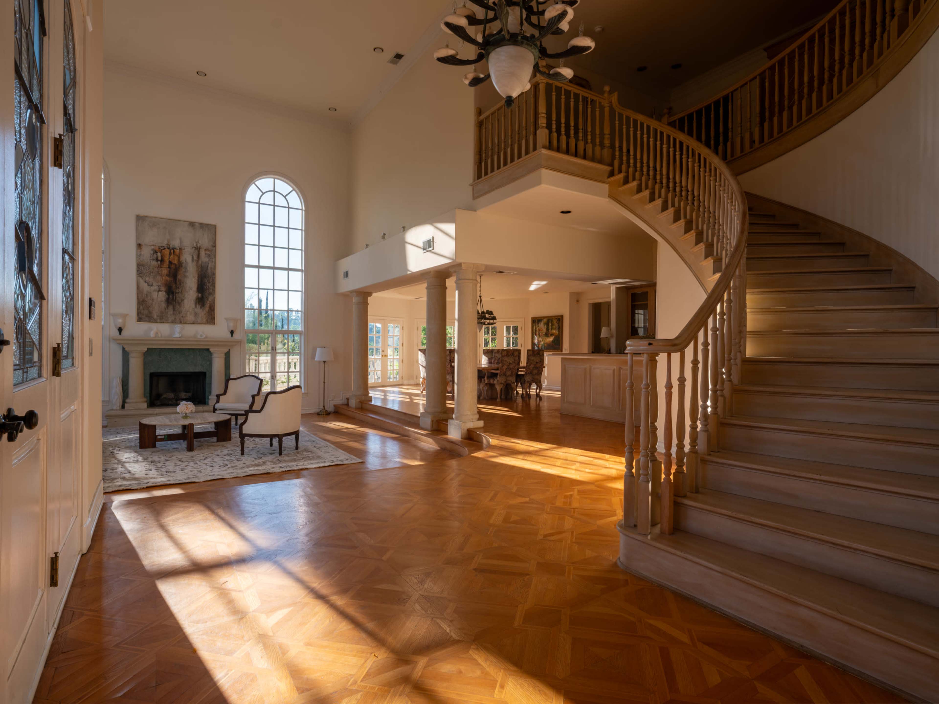 The image shows a spacious foyer with a large staircase, a fireplace, and abundant natural light streaming through tall windows.