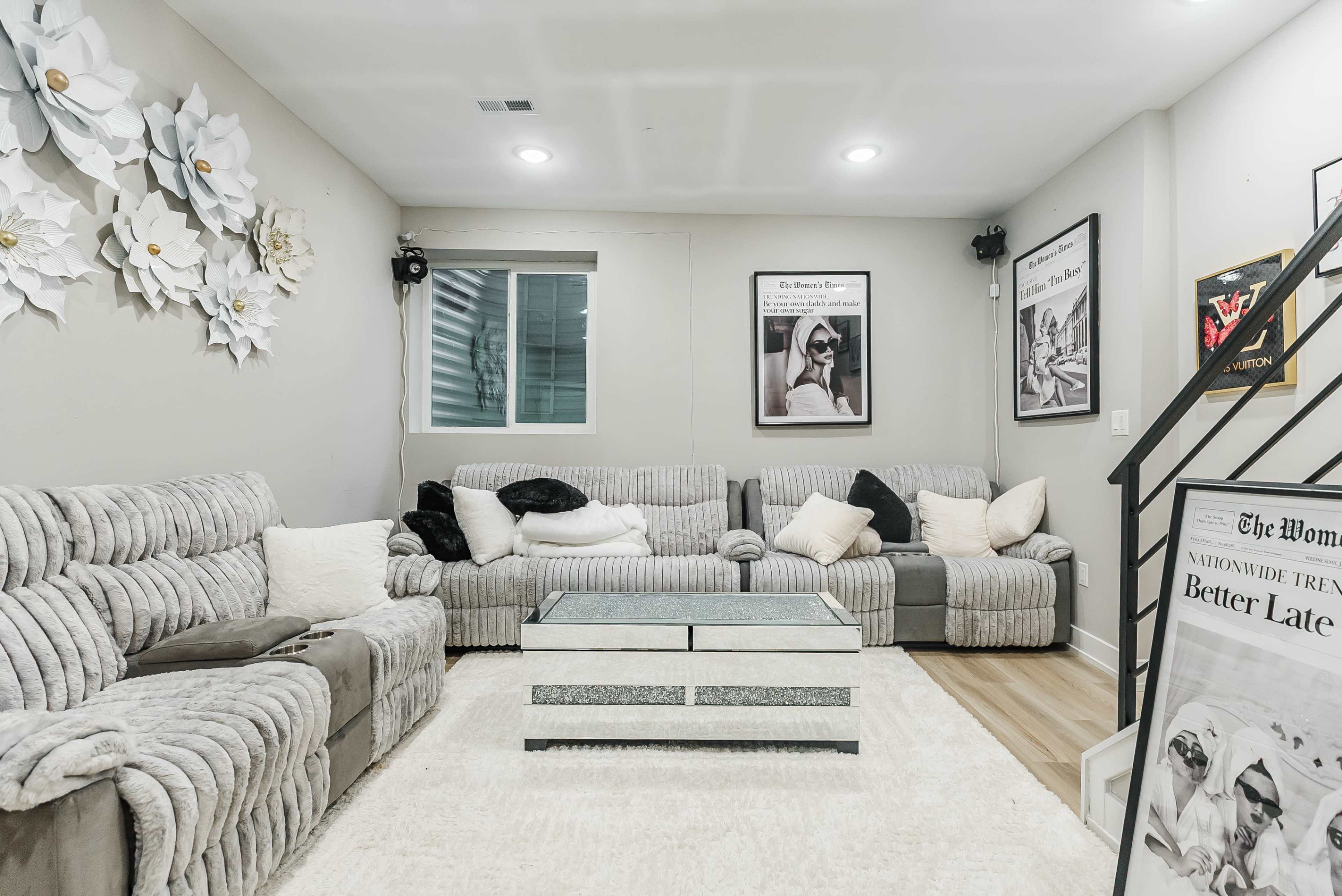 The image shows a modern living room with gray couches, a glass coffee table, and artistic wall decor featuring large paper flowers.