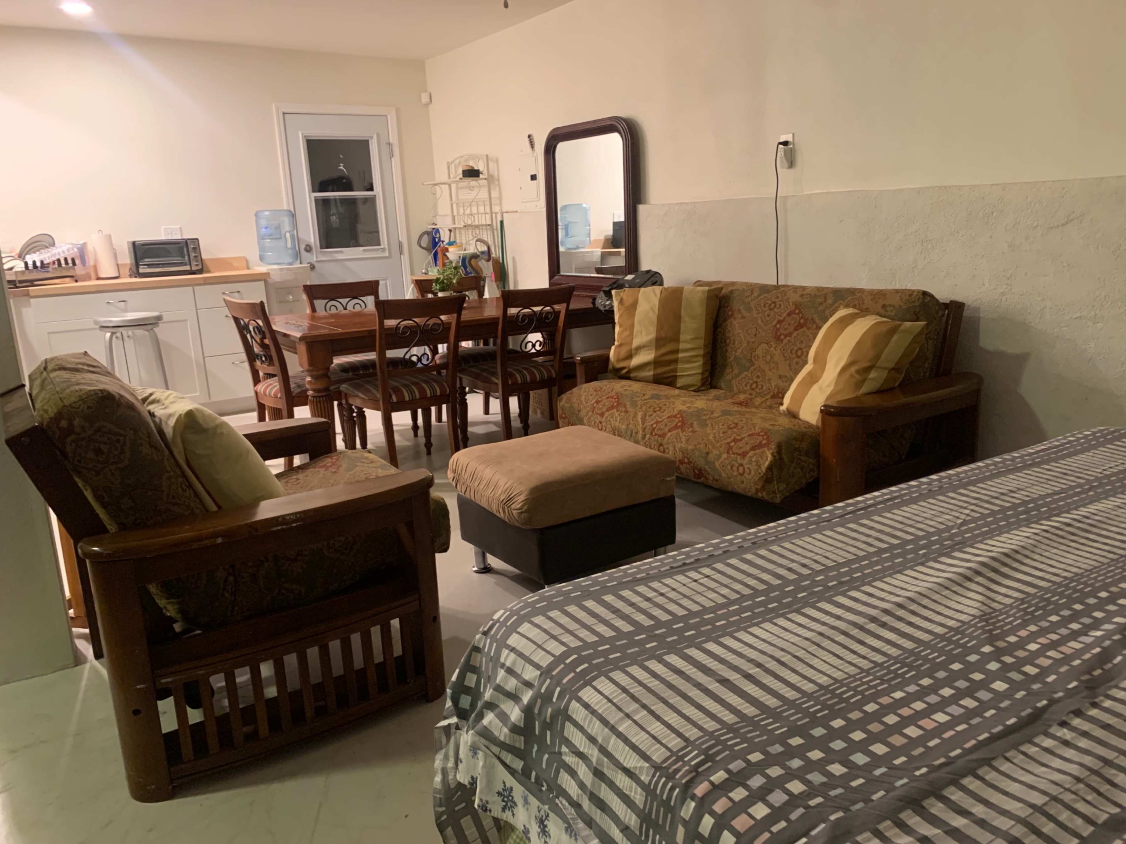 A living space with a small kitchen area, a dining table with six chairs, a sofa set, and a bed with a patterned cover.