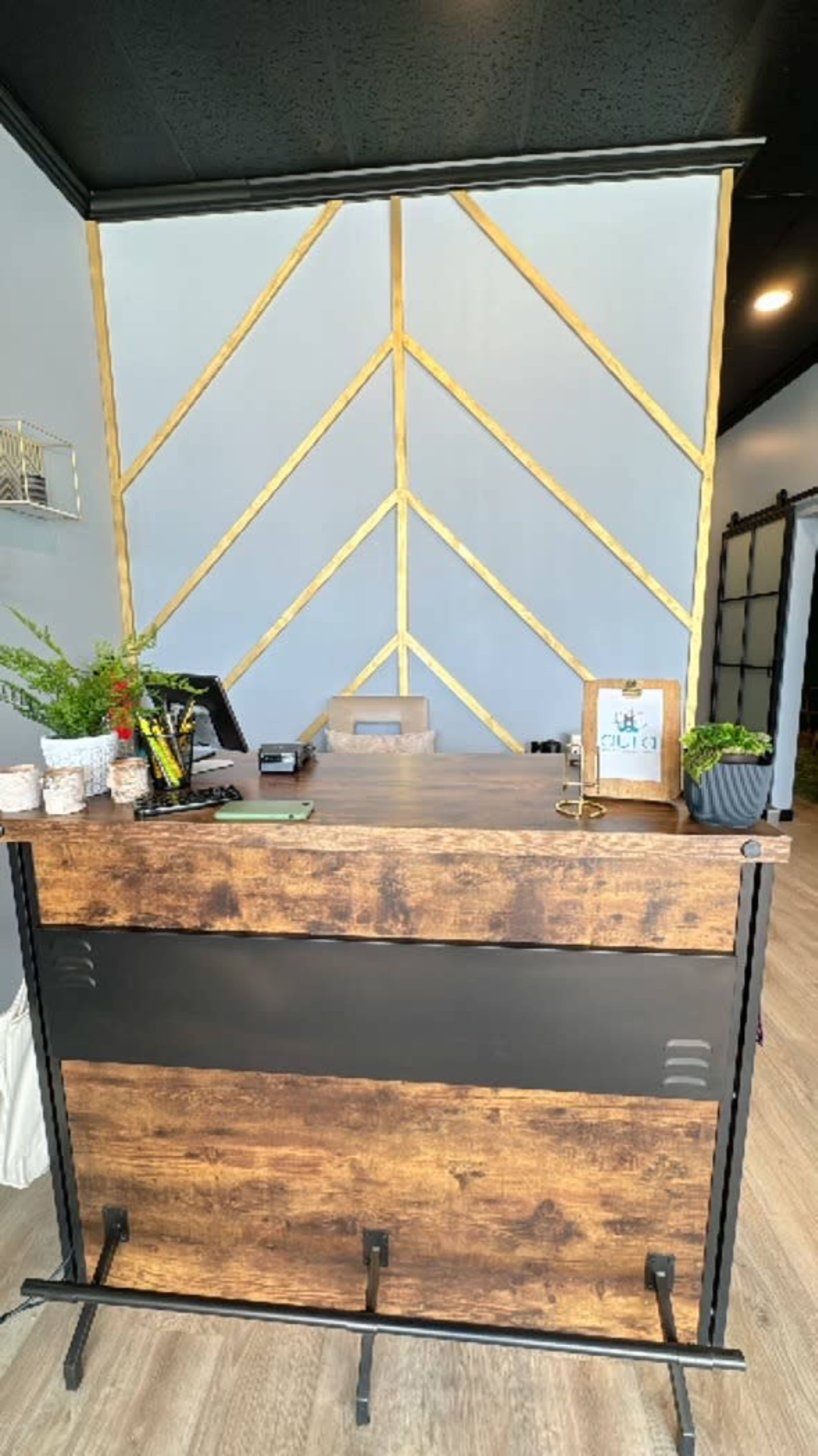 A wooden reception desk is positioned against a geometric accent wall with gold trim, accompanied by plants and decorative items.