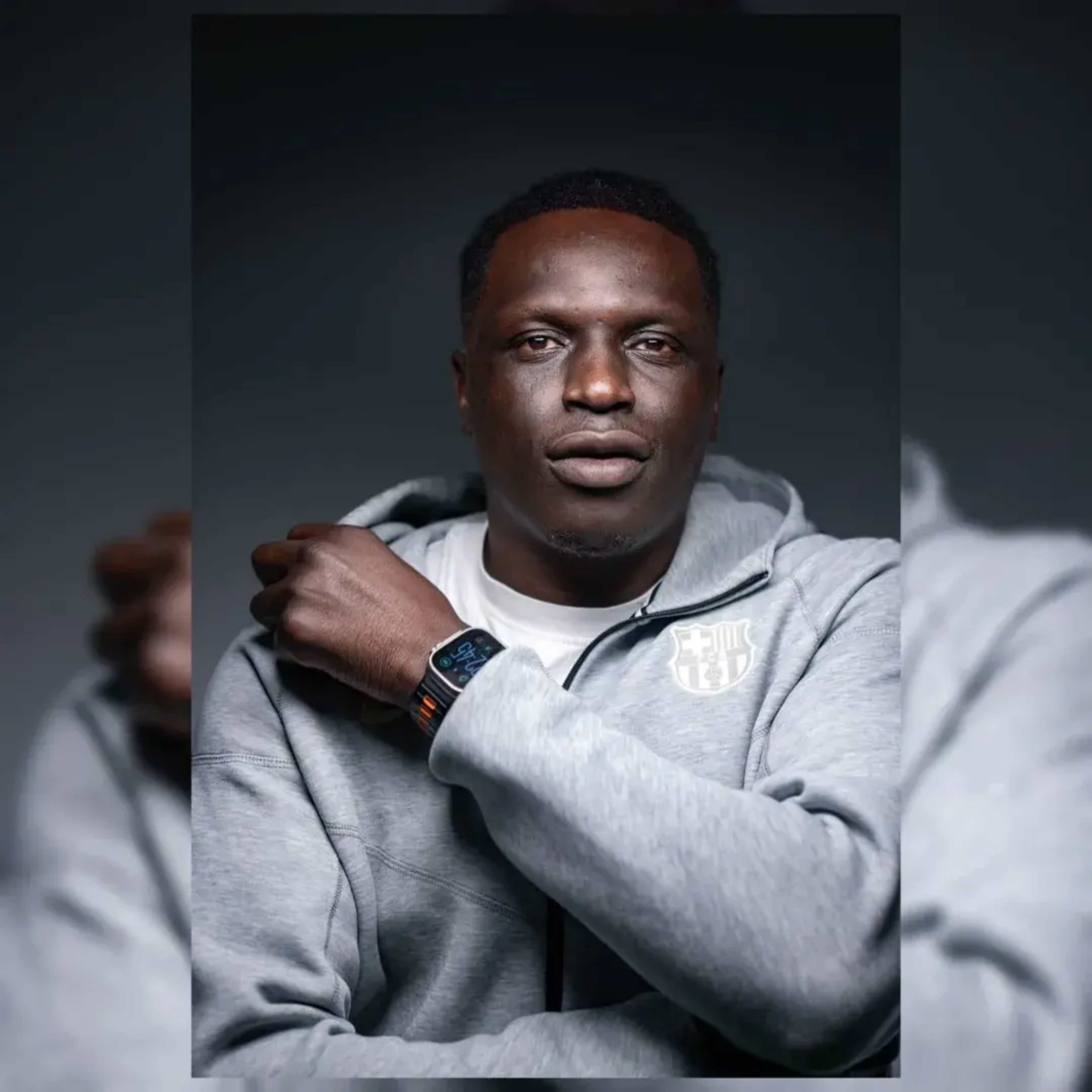 A man wearing a grey hoodie poses with his arm crossed in front of him, showcasing a smartwatch on his wrist.