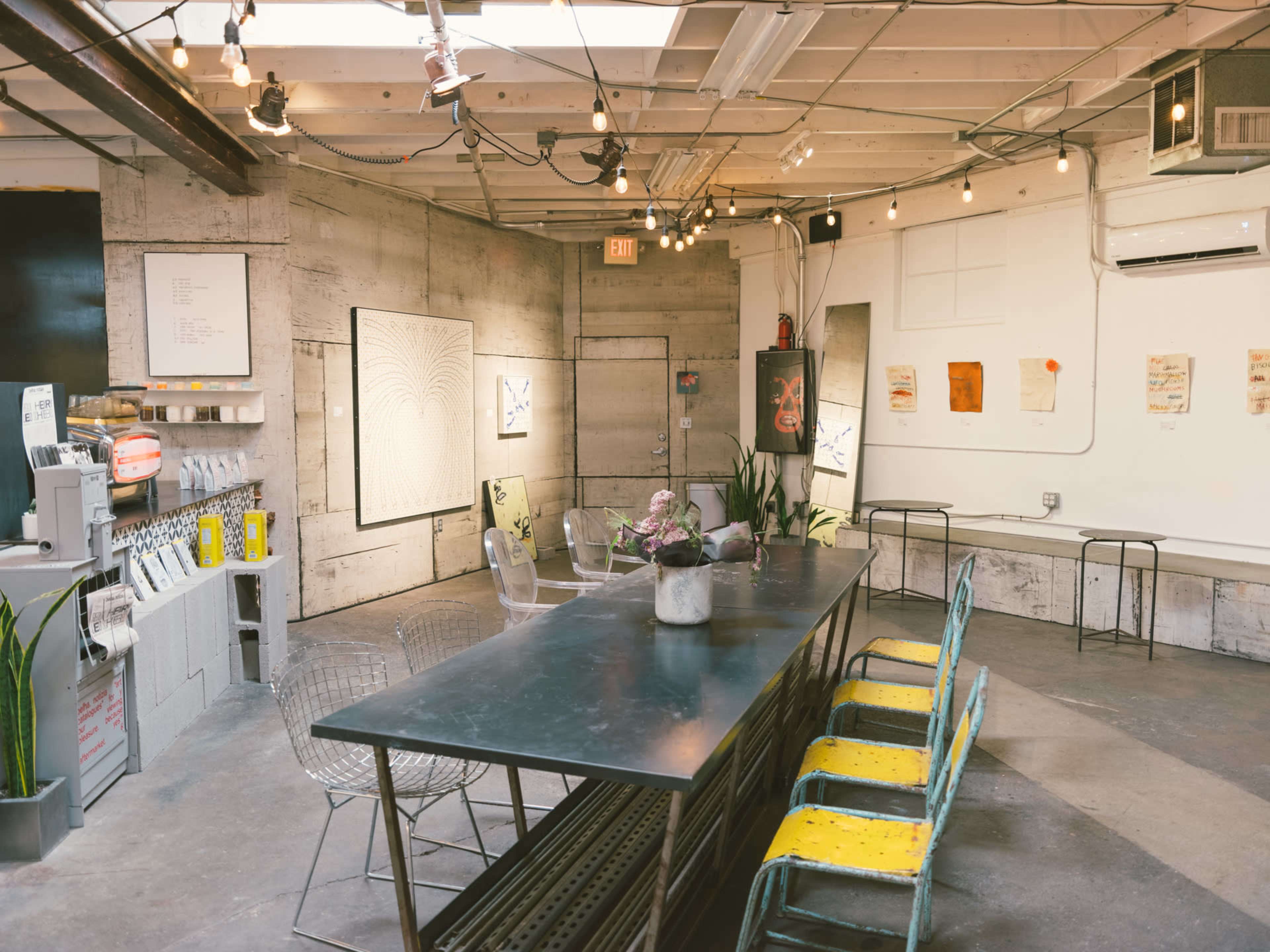 The image shows a modern, industrial-style interior with a long metal table surrounded by mismatched chairs, illuminated by hanging light bulbs.