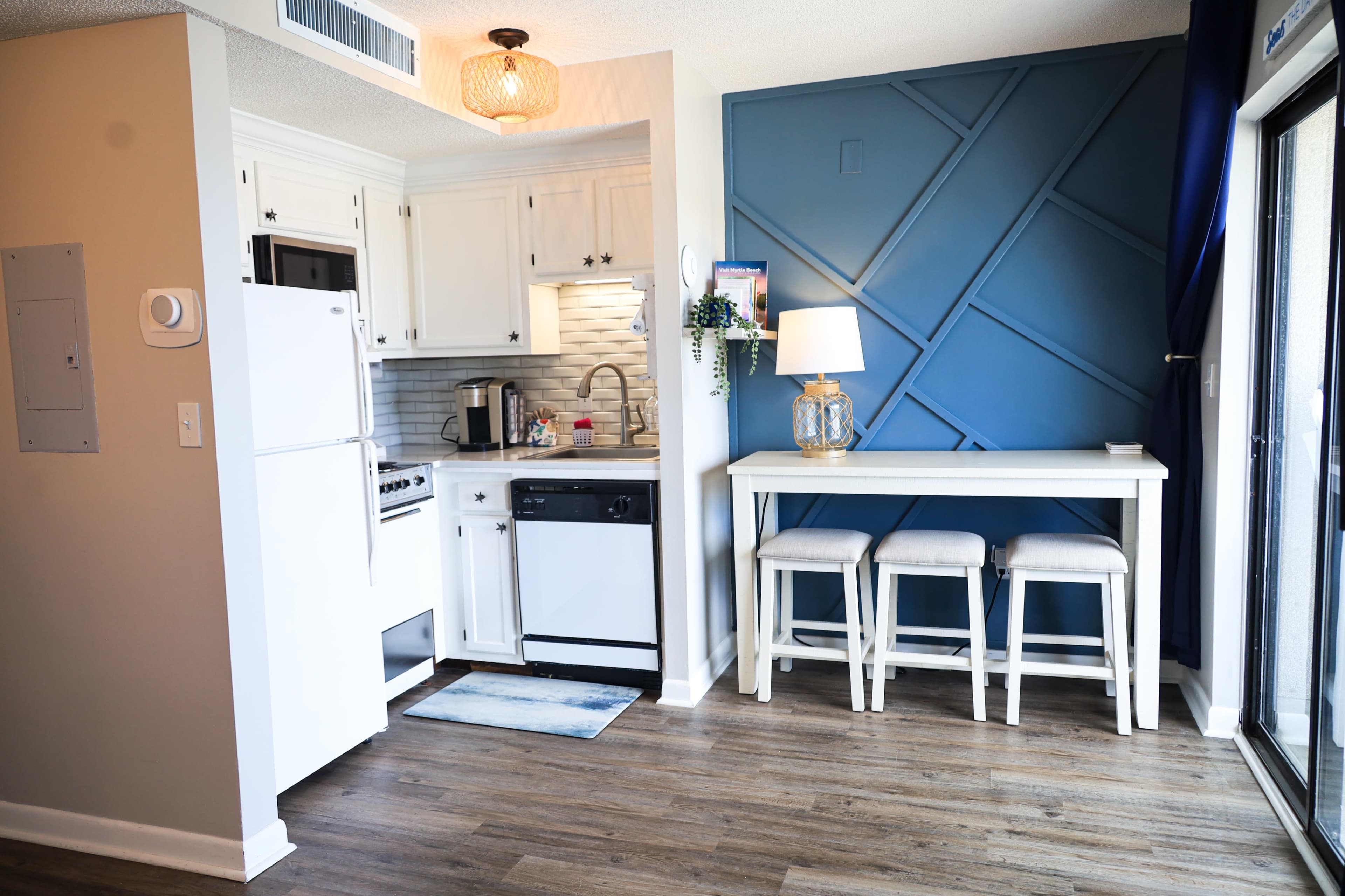 A modern kitchen and dining area feature white cabinetry, a blue accent wall, and a set of three stools at a small table.