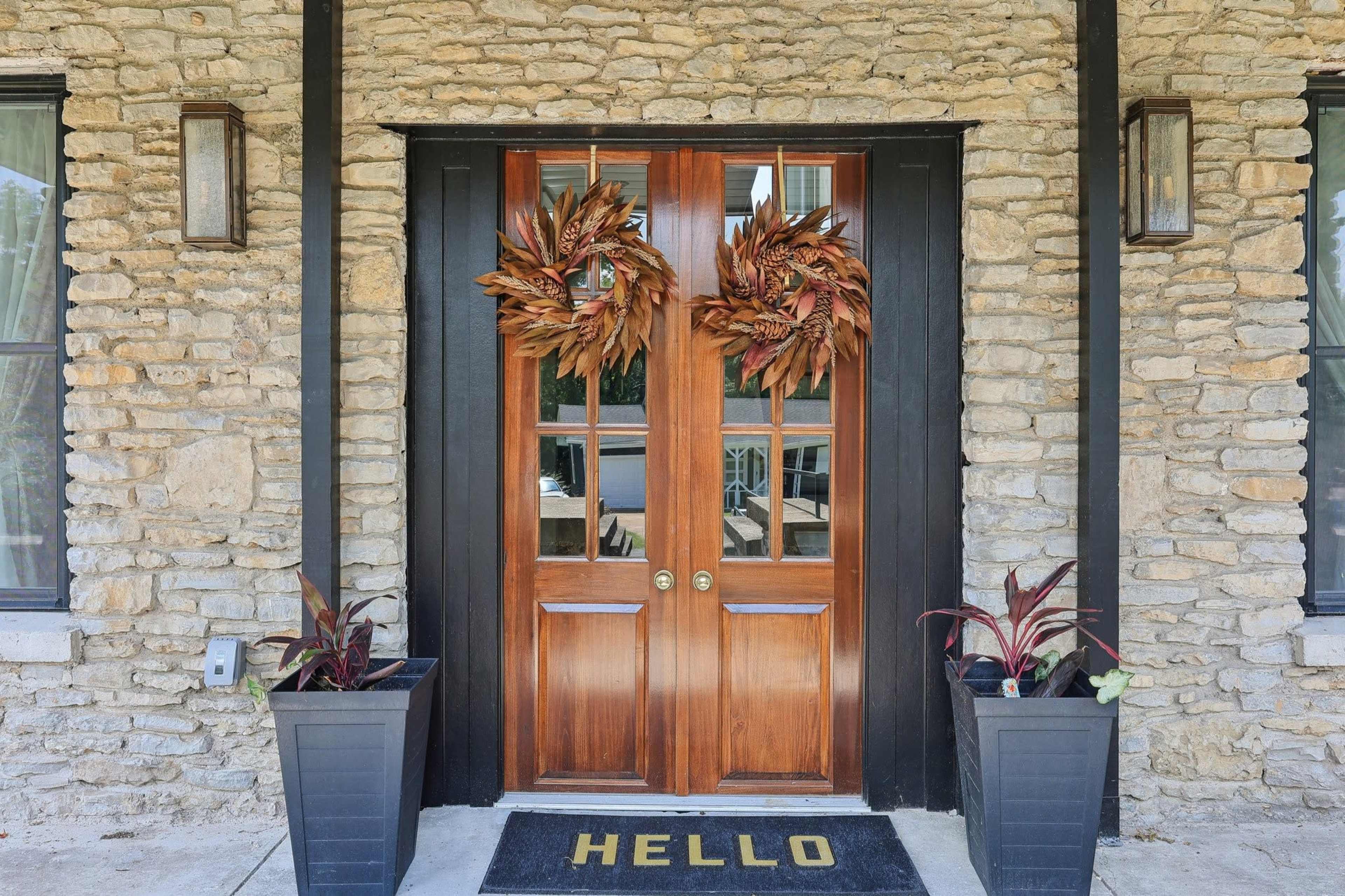 A pair of wooden double doors with glass panels, flanked by decorative wreaths, and surrounded by potted plants and a welcome mat.