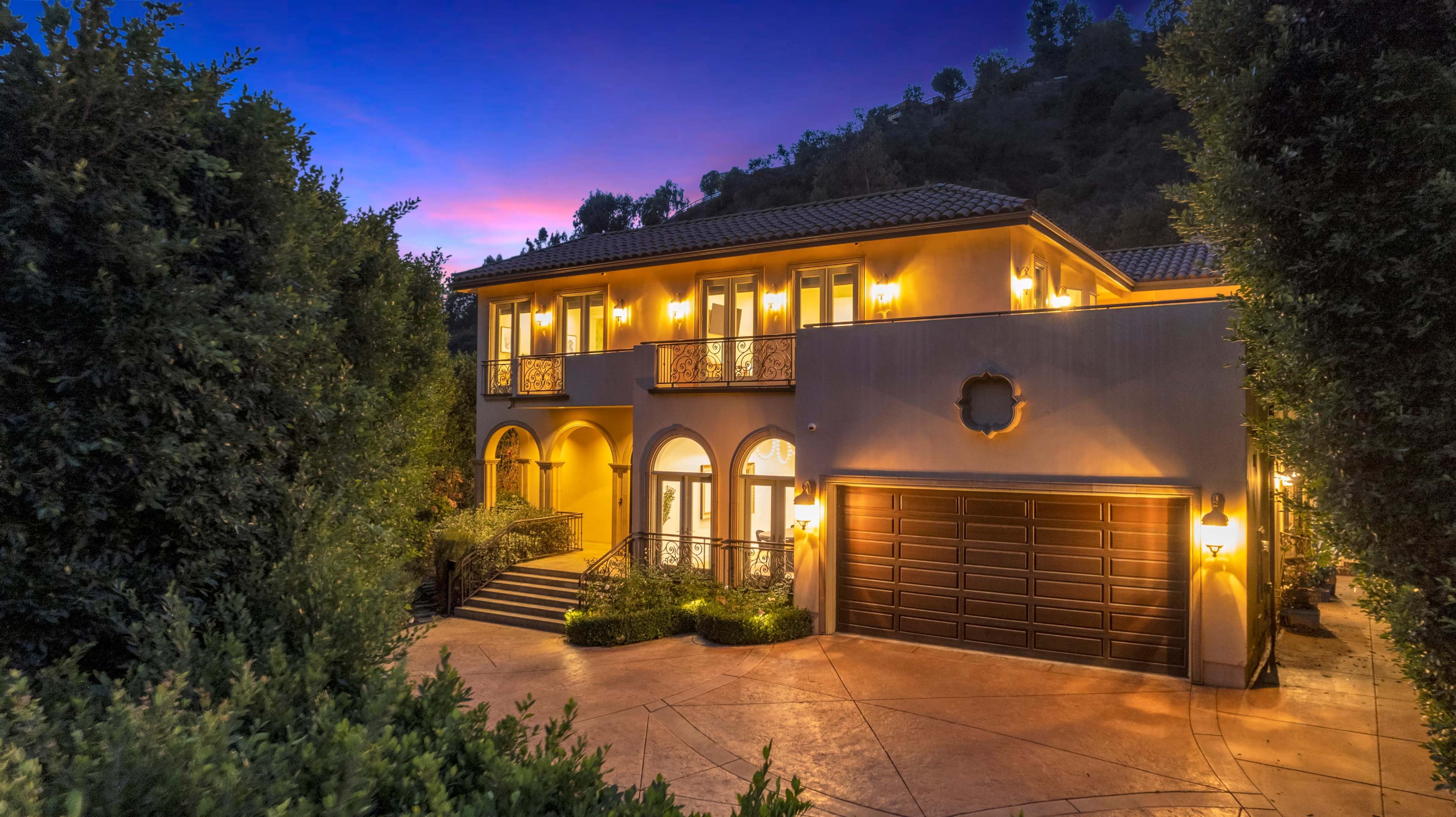 A two-story house with a tile roof and a spacious driveway is illuminated by outdoor lighting at dusk, surrounded by manicured greenery.
