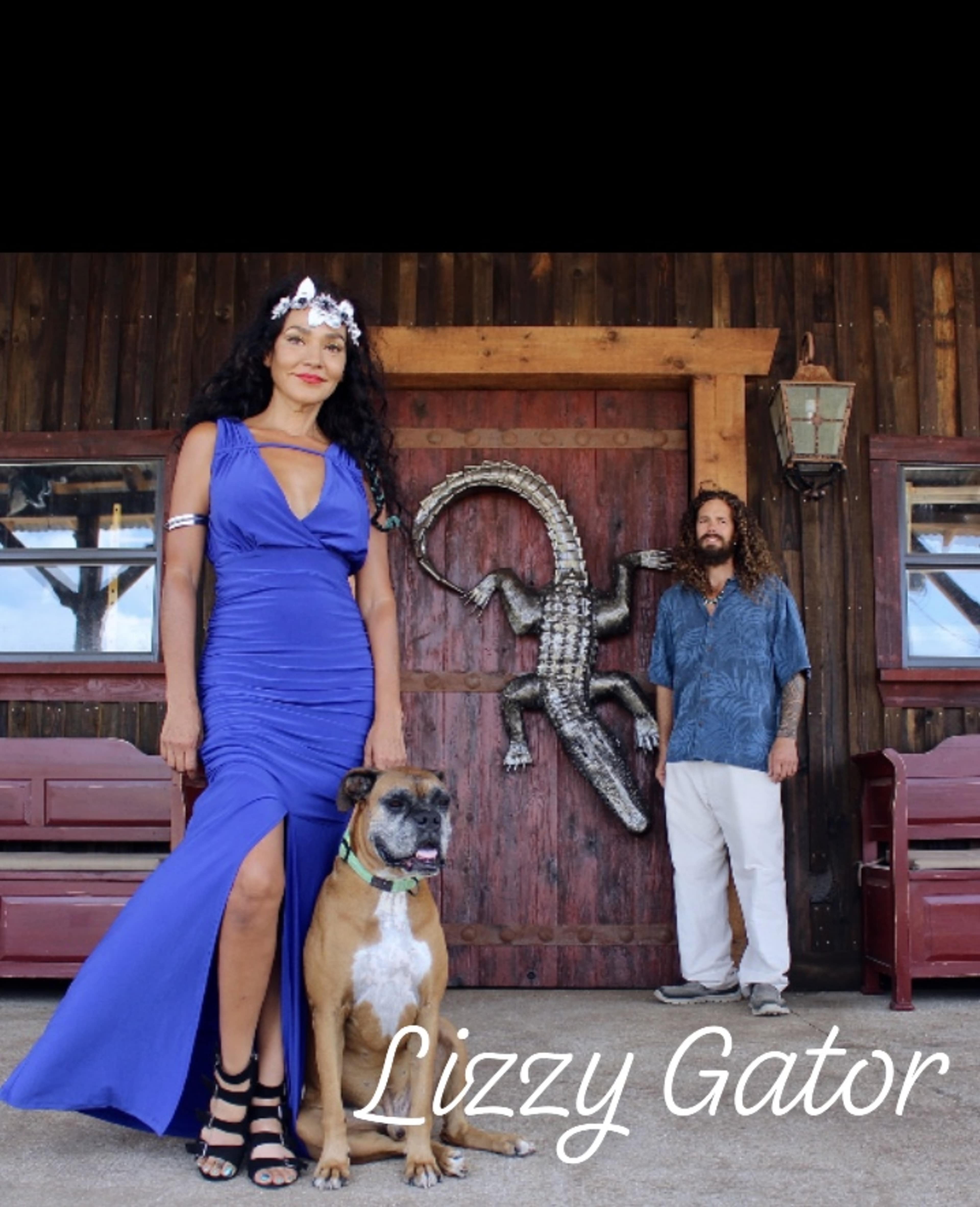 A woman in a blue dress stands next to a dog, while a man in a patterned shirt stands behind them in front of a wooden structure featuring a decorative alligator.