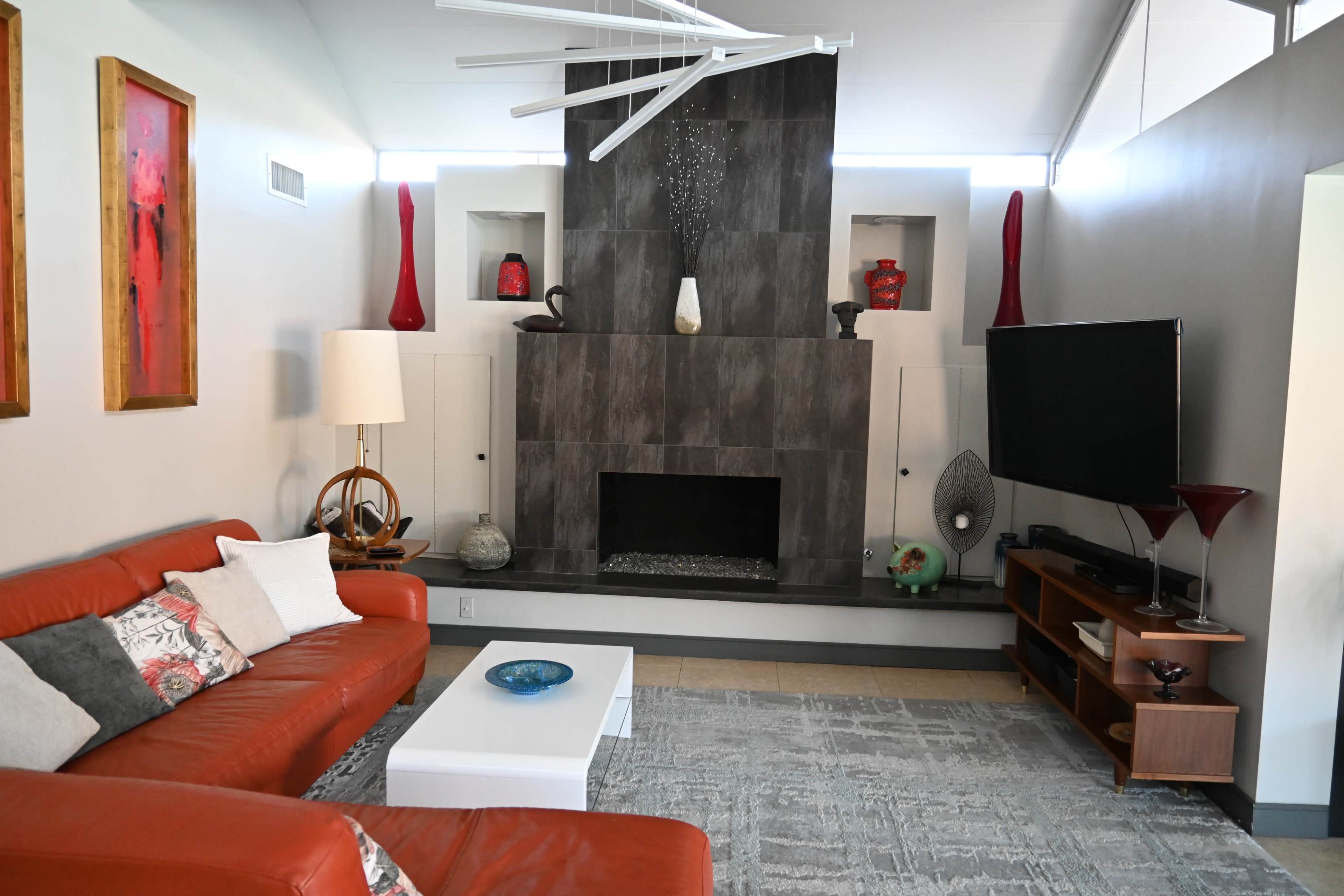 The image shows a modern living room featuring a red leather sofa, a white coffee table, a gray stone fireplace, and decorative vases on a shelf.